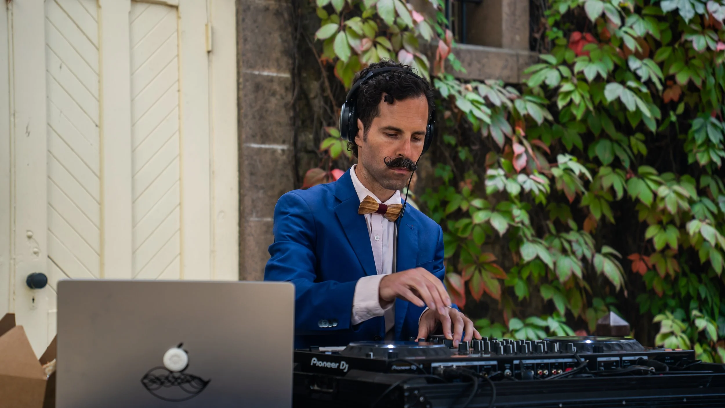 A man dressed in a blue suit with a wooden bow tie is DJing outdoors, wearing large headphones and focusing on the mixing console in front of him. There are plants and a house door in the background.