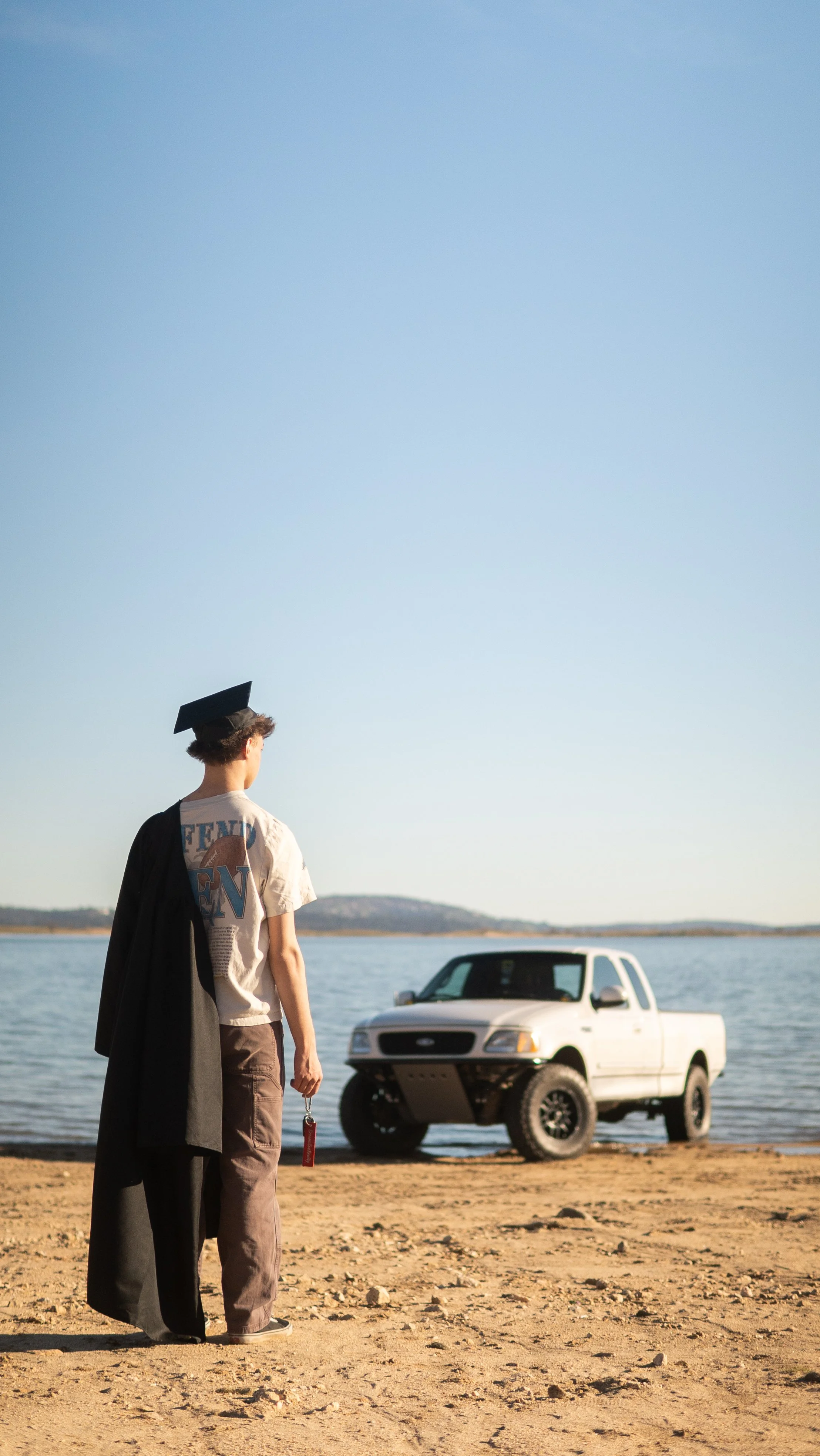 A young man with a graduation cap on his head stands on a beach near water, holding a set of keys, with a white pickup truck parked nearby, during daytime.