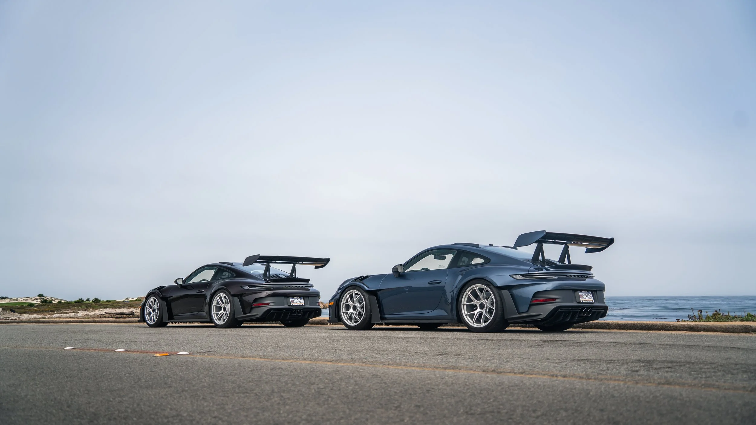 Two black luxury sports cars with large rear spoilers parked side by side near the ocean under a cloudy sky.