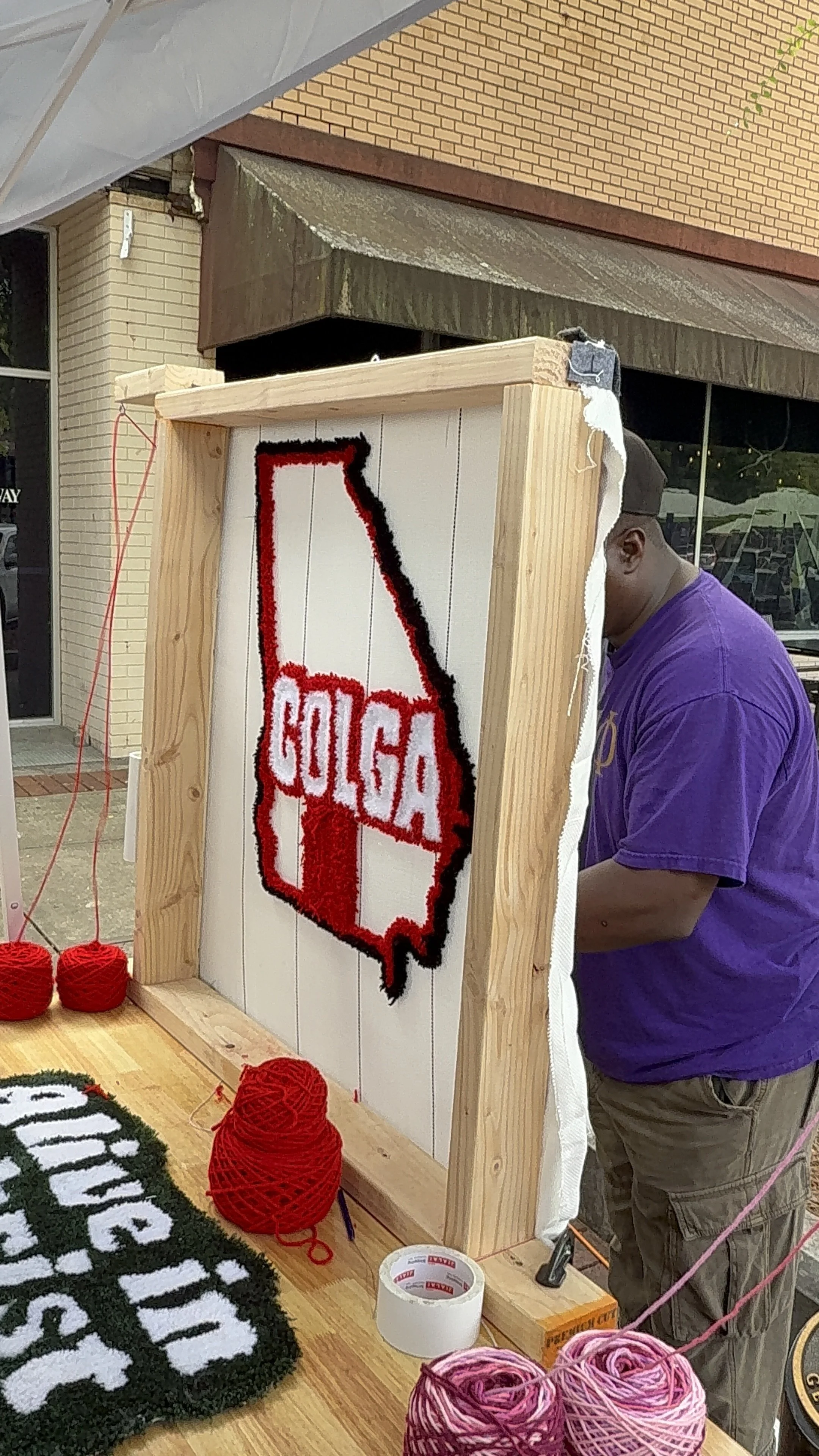 Yarn craft booth displaying a Texas-shaped sign with the word 'GOLGA' and red, black, and white yarn accents, with yarn balls and craft supplies on the table.