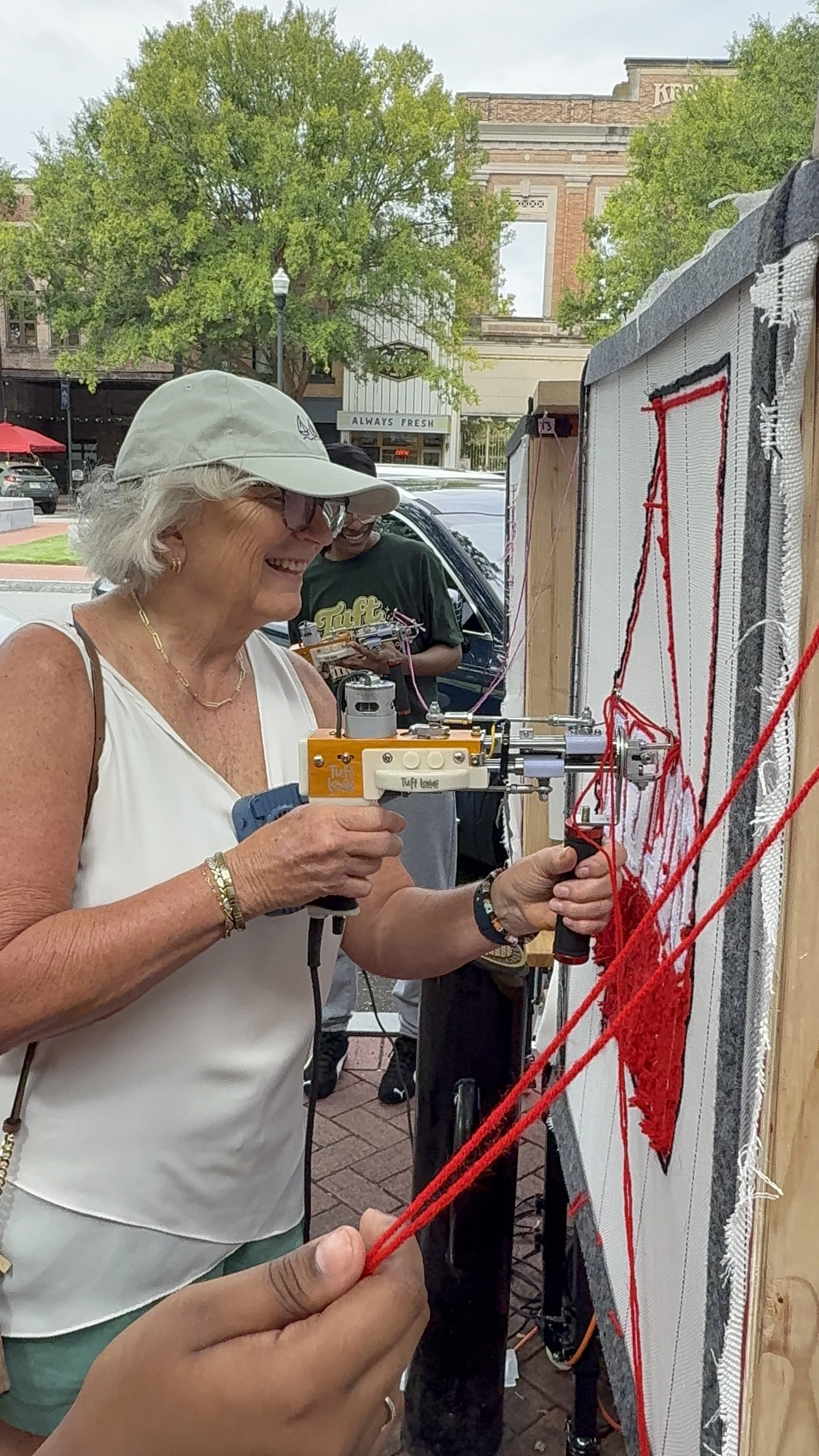 A woman with white hair, sunglasses, and a white hat smile as she uses a small machine to sew red yarn onto a white fabric with red thread. Several people stand nearby, and a brick building with trees and a sign reading 'ALWAYS FRESH' are in the back