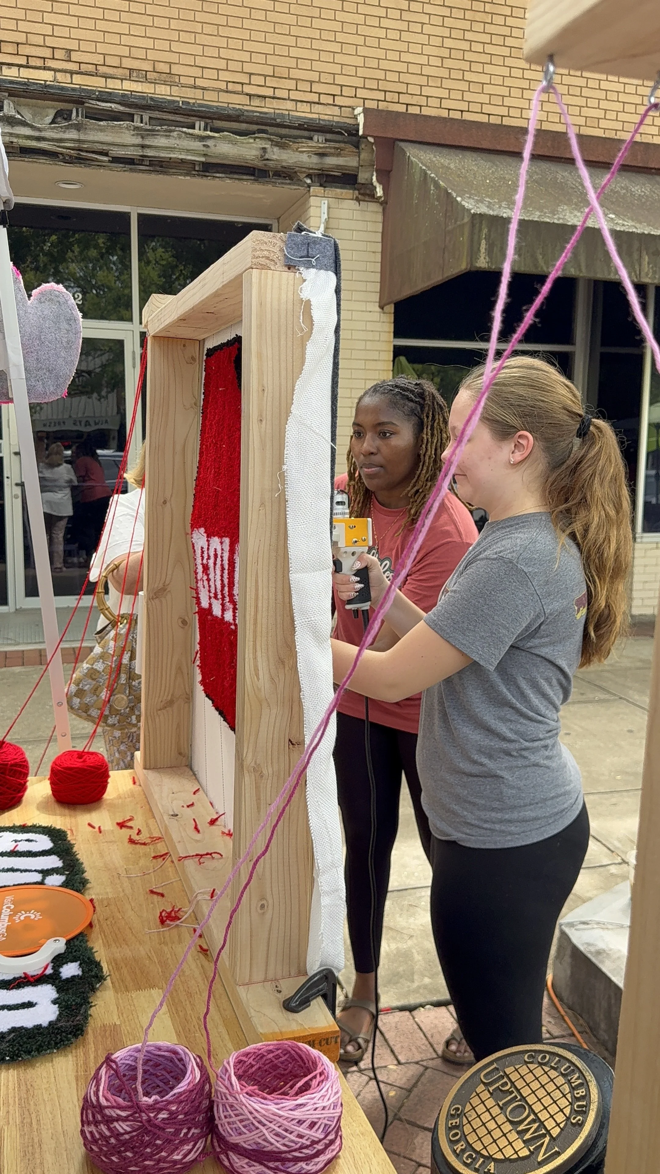 Two women look at yarn and crochet projects on a wooden display at an outdoor event. The display has balls of yarn, some partially crocheted items, and a framed rectangular piece they are examining. The background shows a brick building and a window 