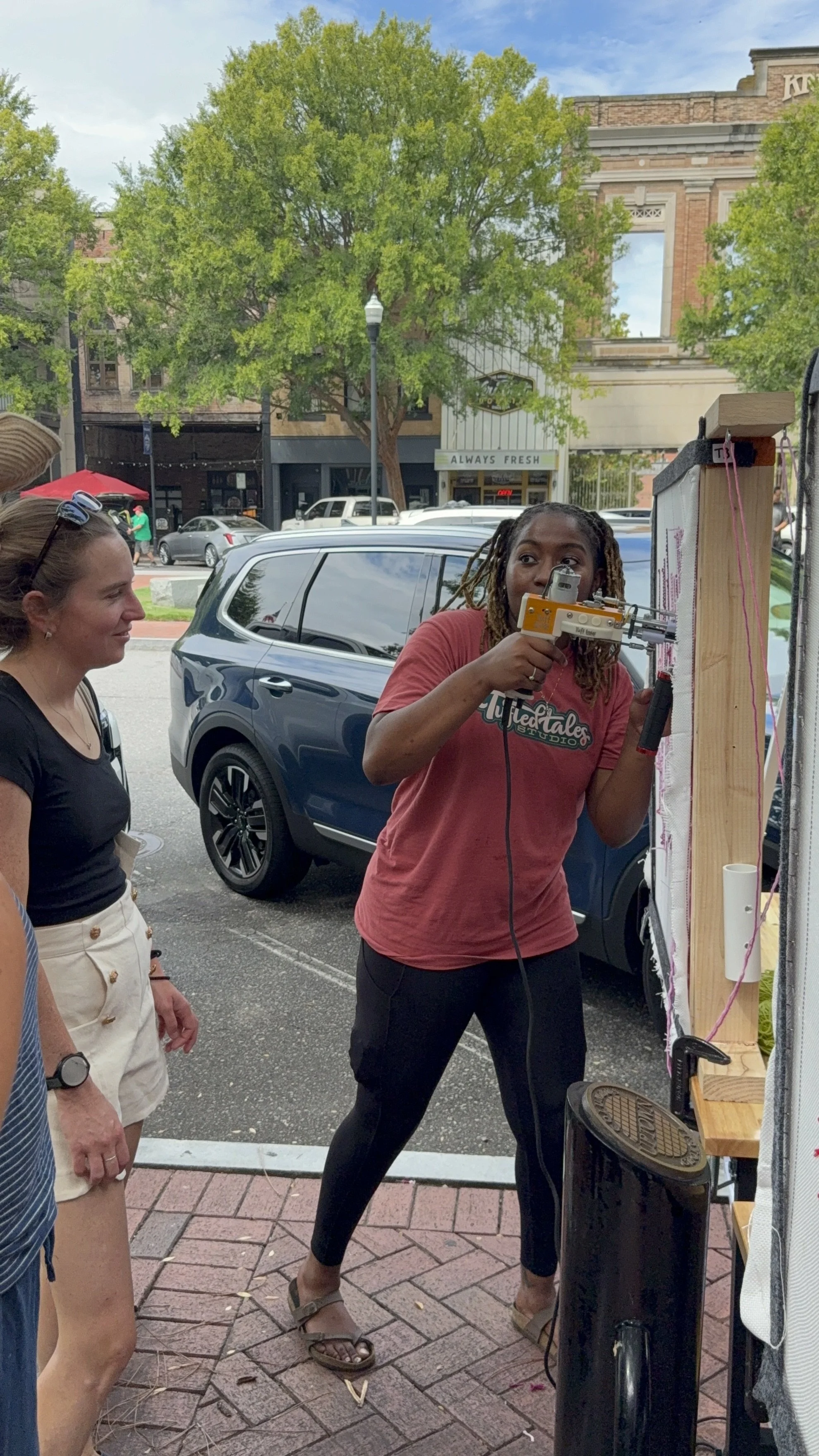 A woman with dreadlocks using a hot glue gun on a craft project outdoors, with a woman in a black top and white shorts watching. There are cars parked behind them, and trees and a building with storefronts in the background.