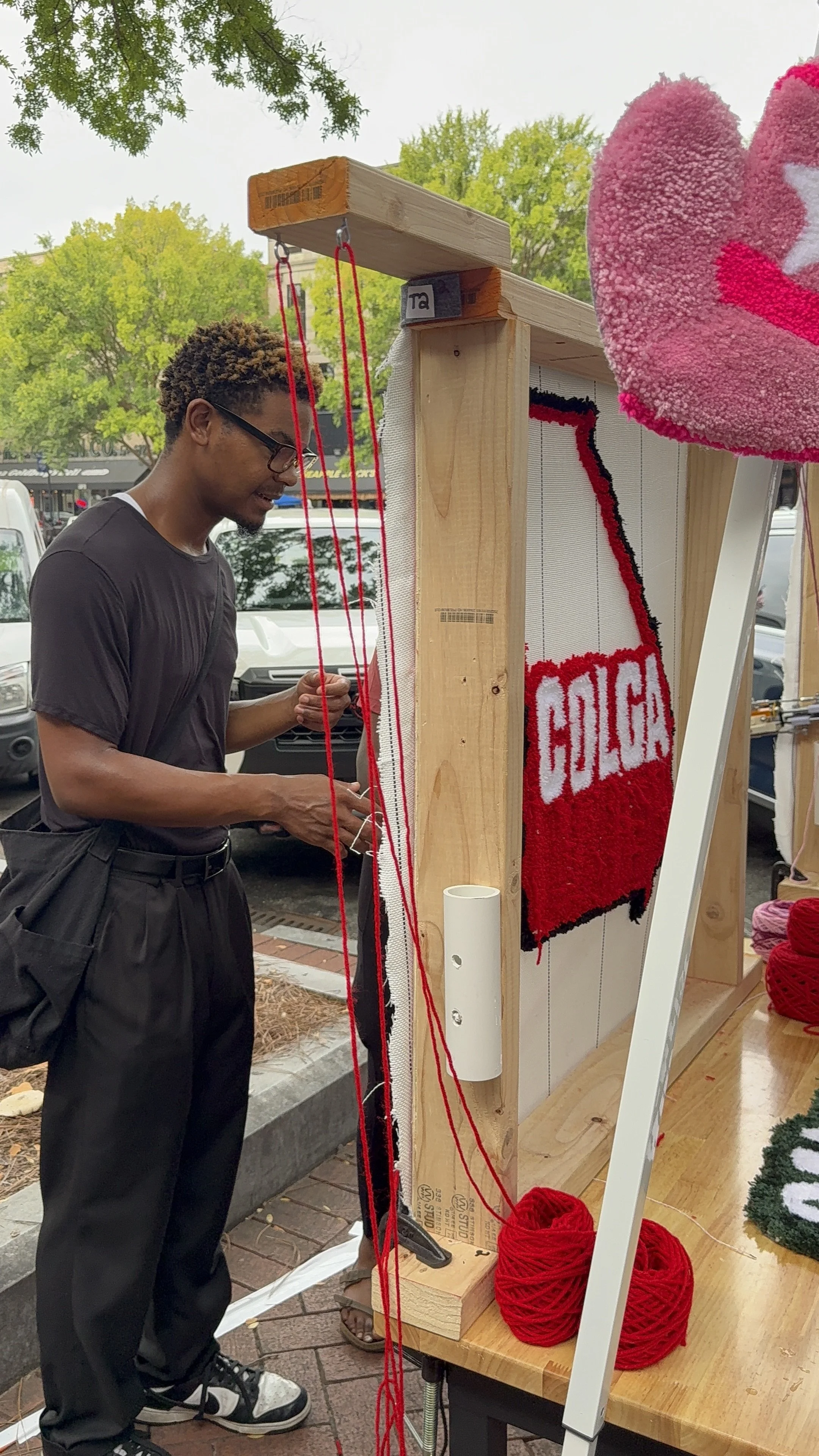 A person with glasses and dark clothing knitting or working on a craft project at an outdoor setup. Yarn and a partially completed red and white quilt or tapestry with the word 'GOLGA' are visible.