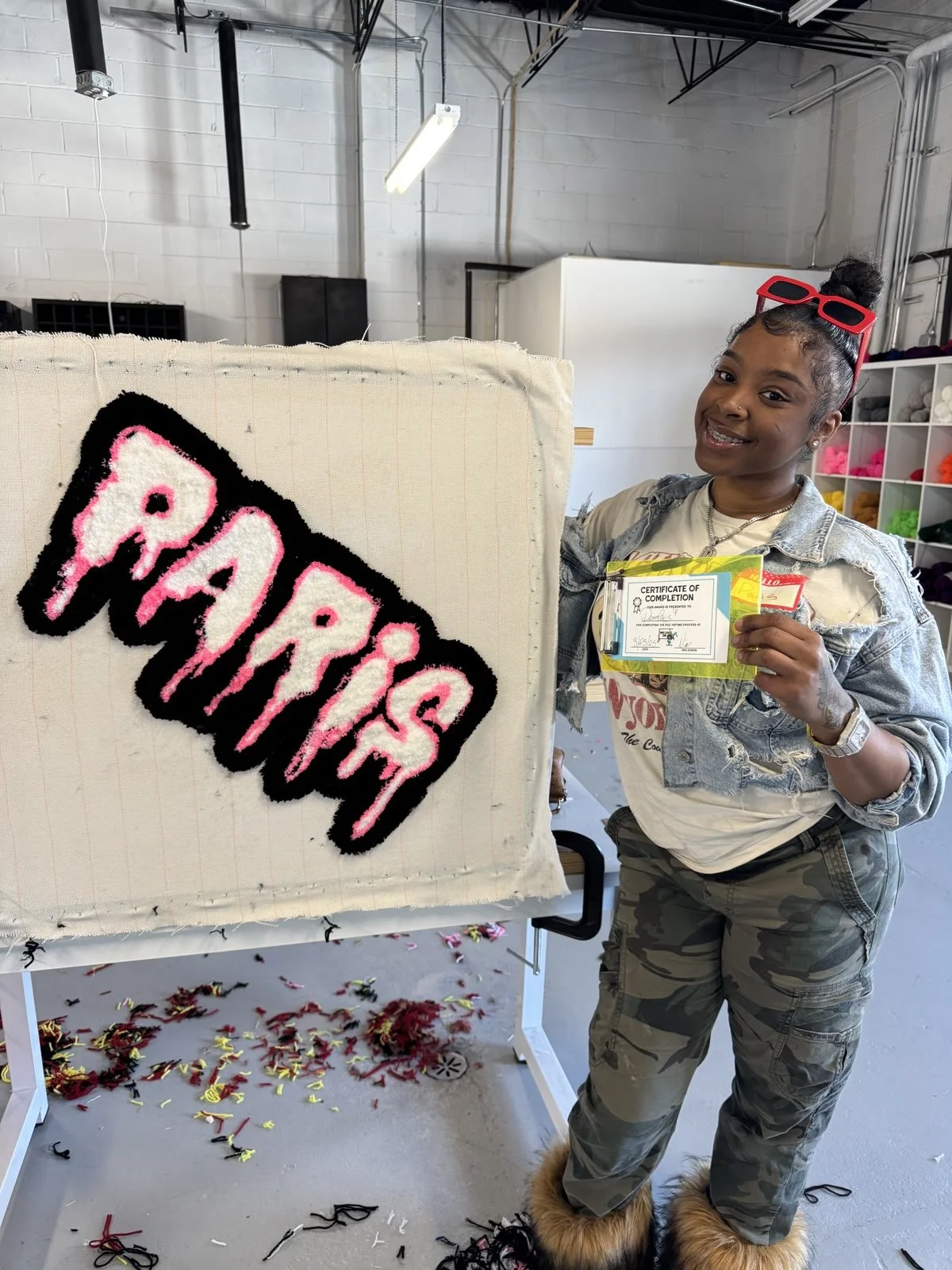 A woman with dark hair in a bun and a nose piercing holds a certificate of completion and stands next to a partially completed yarn art piece with the word 'PARADISE' in pink and black. She is smiling and wearing sunglasses on her head, a light-color