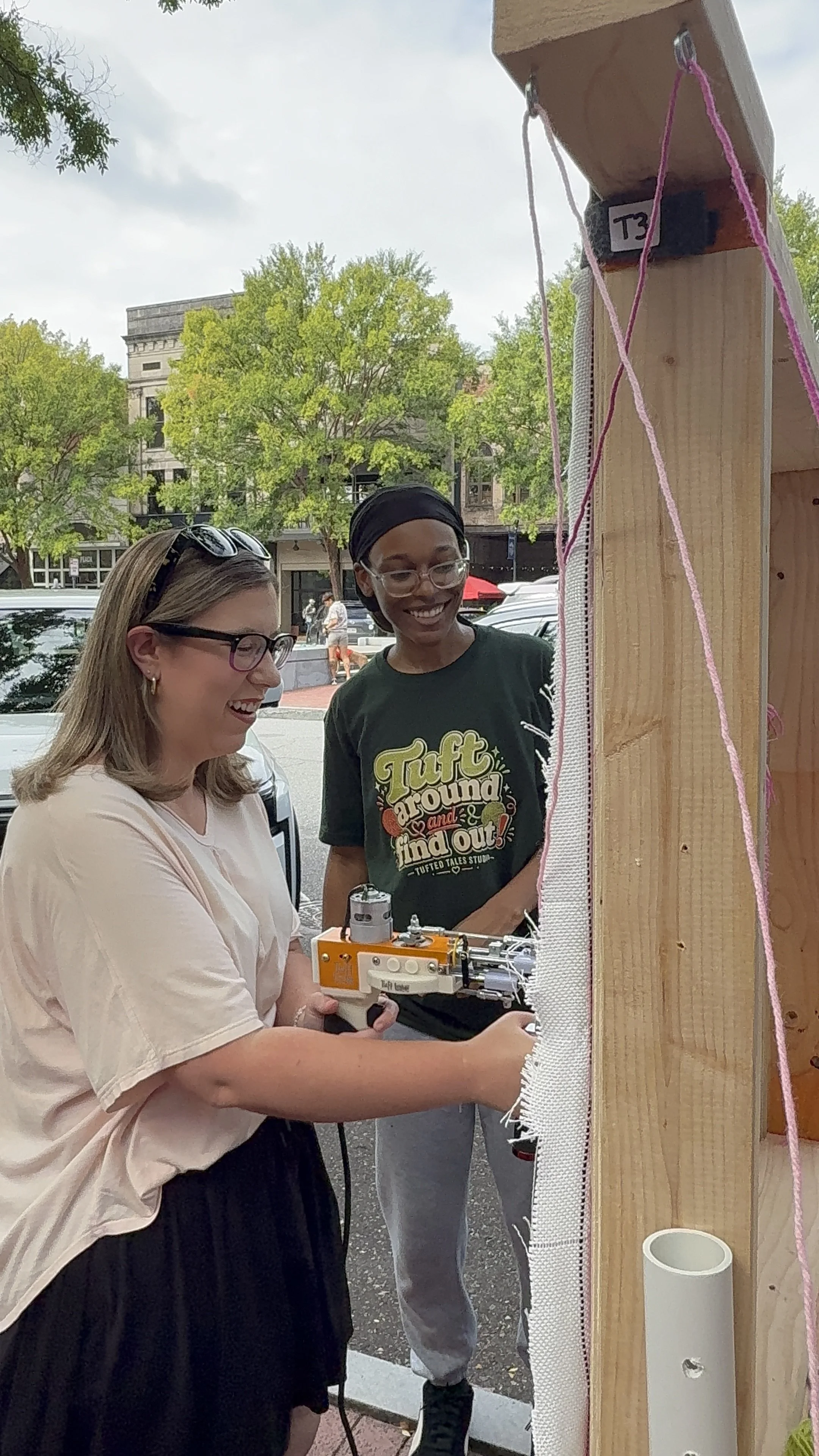 Two women and a young man are smiling and working together on a project outdoors, with trees and a building in the background. They appear to be engaged with a robotics or engineering project.