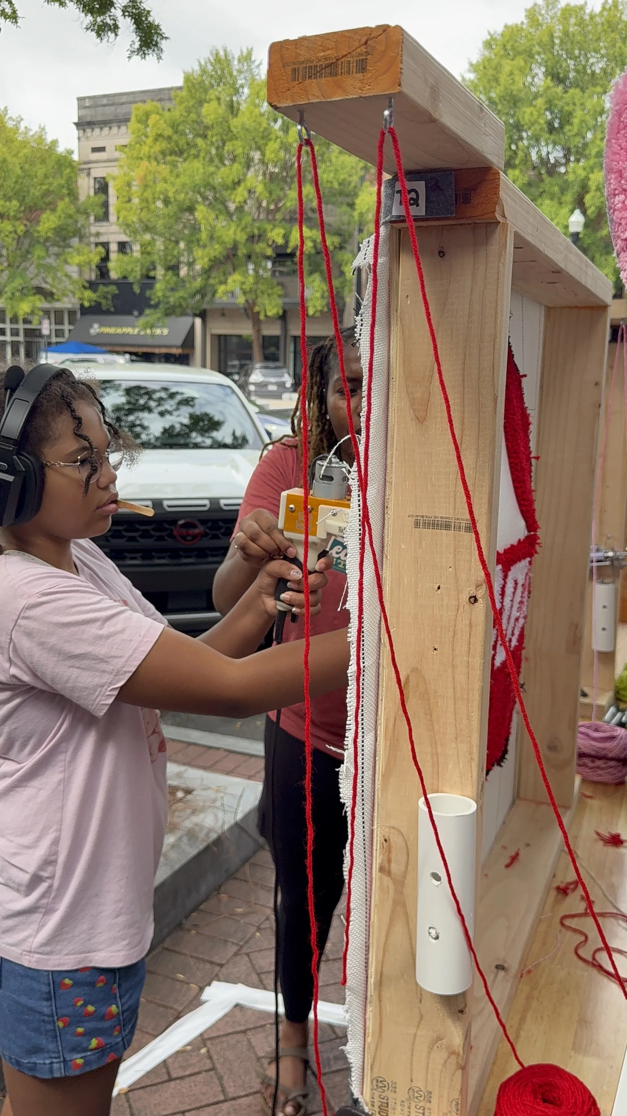 Two women working on a craft project with a vertical wooden structure, yarn, and a sewing machine outdoors in a city setting.