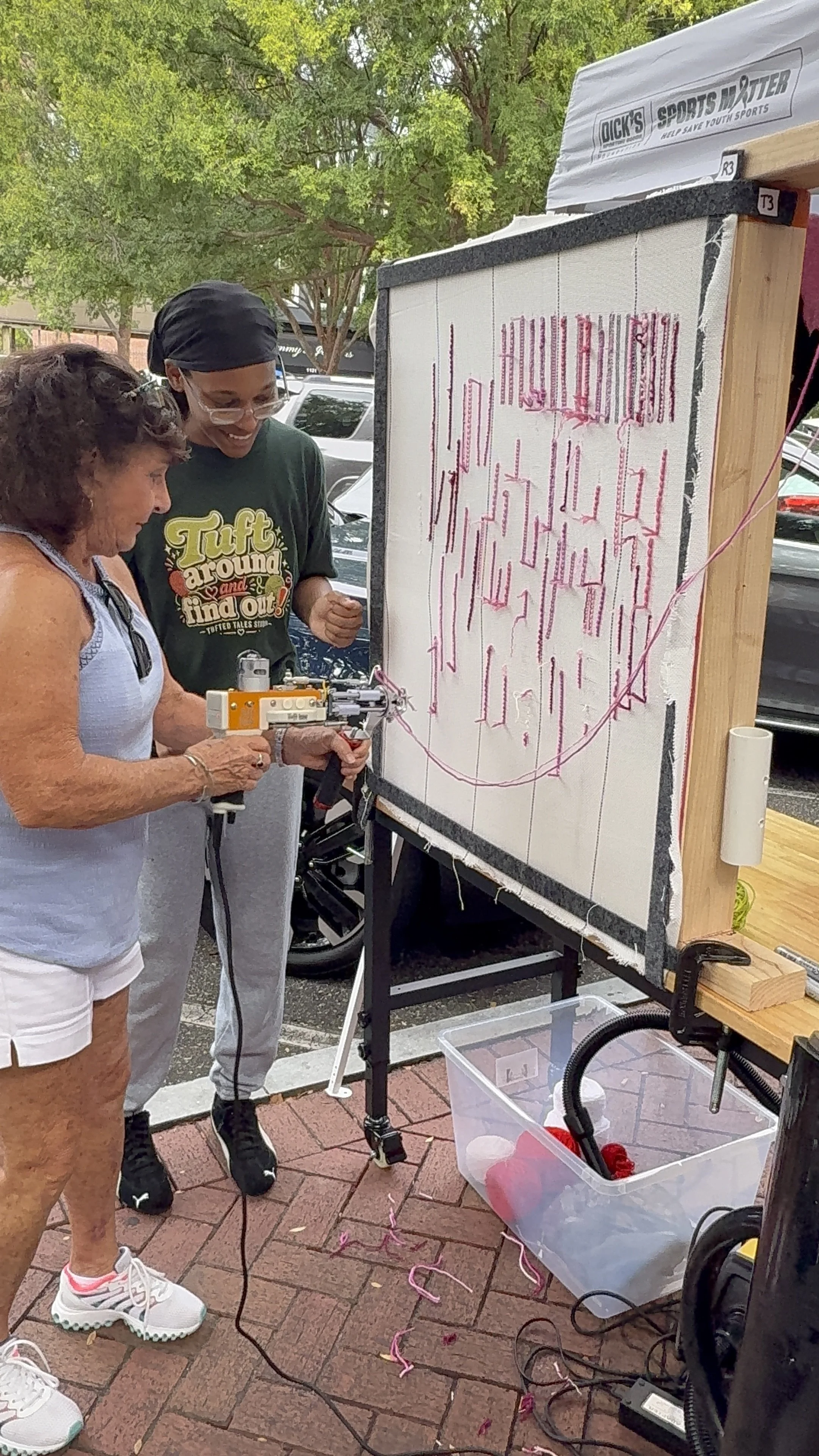 Two women are creating a large, colorful piece of thread art on a fabric canvas, which is mounted on a wooden frame outdoors. One woman, older, is holding a device that controls the thread, while the other, younger, is observing and smiling. There ar