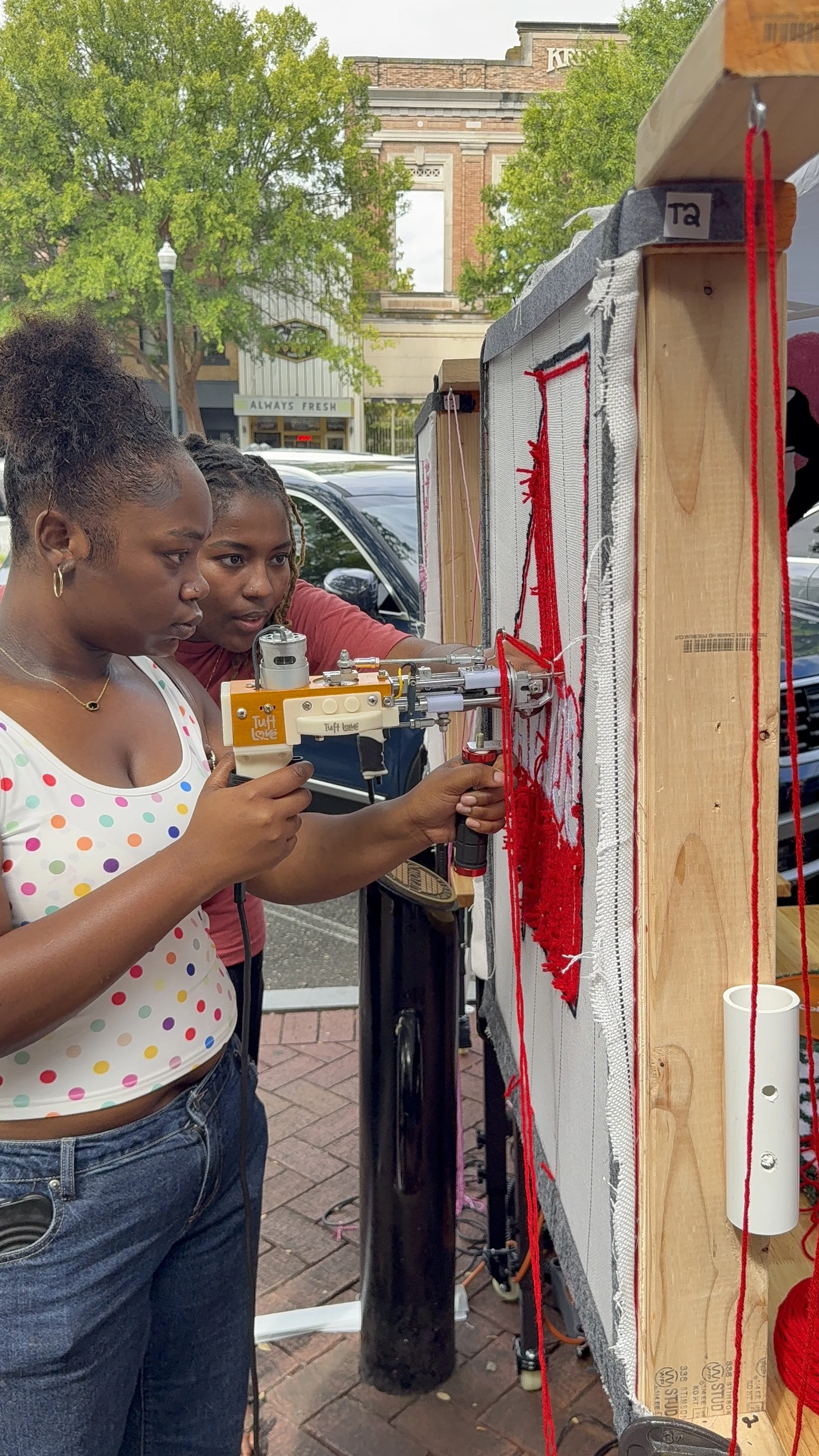 Two women working on a puppet show, using a small mechanical puppet on a string puppet stage outdoors. The background has trees, cars, and a building with a sign that says 'Always Fresh'.