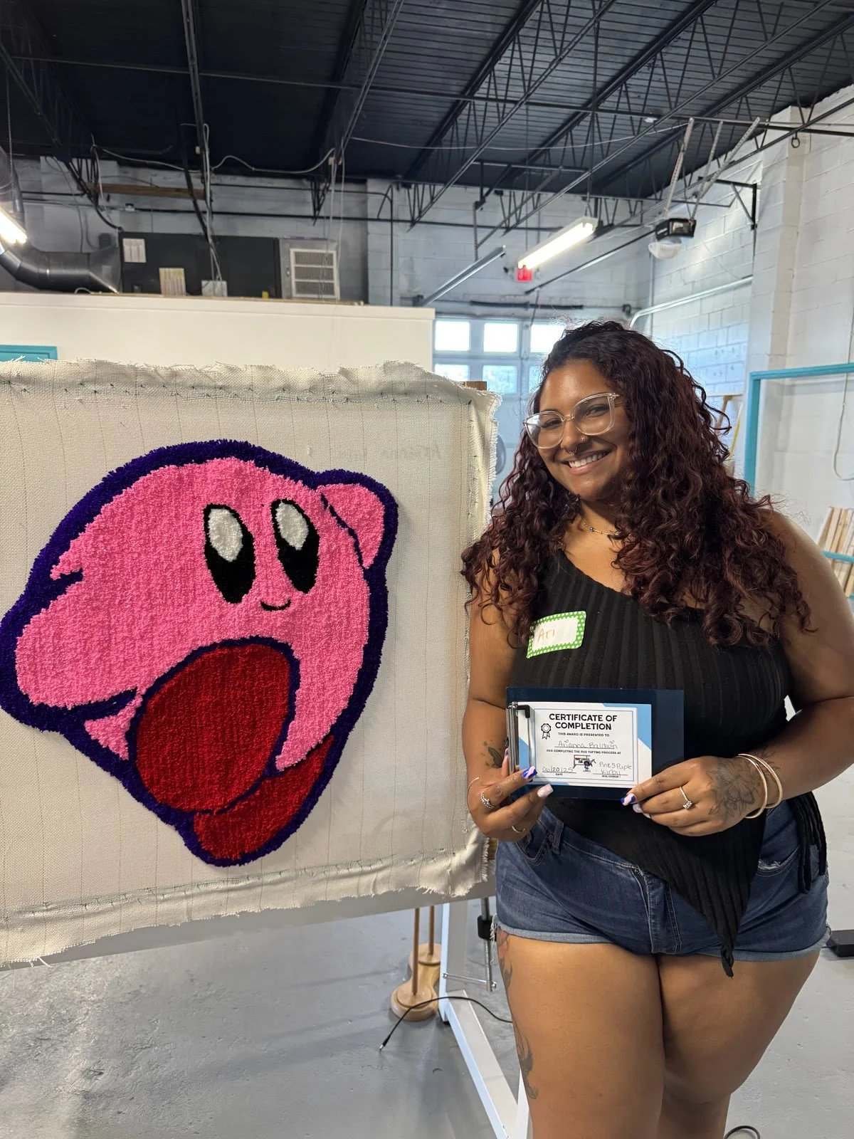 A woman with long curly hair, glasses, and a black sleeveless top holding a certificate of completion, standing next to a display of a pink Kirby character made of textured material inside an industrial-style building.