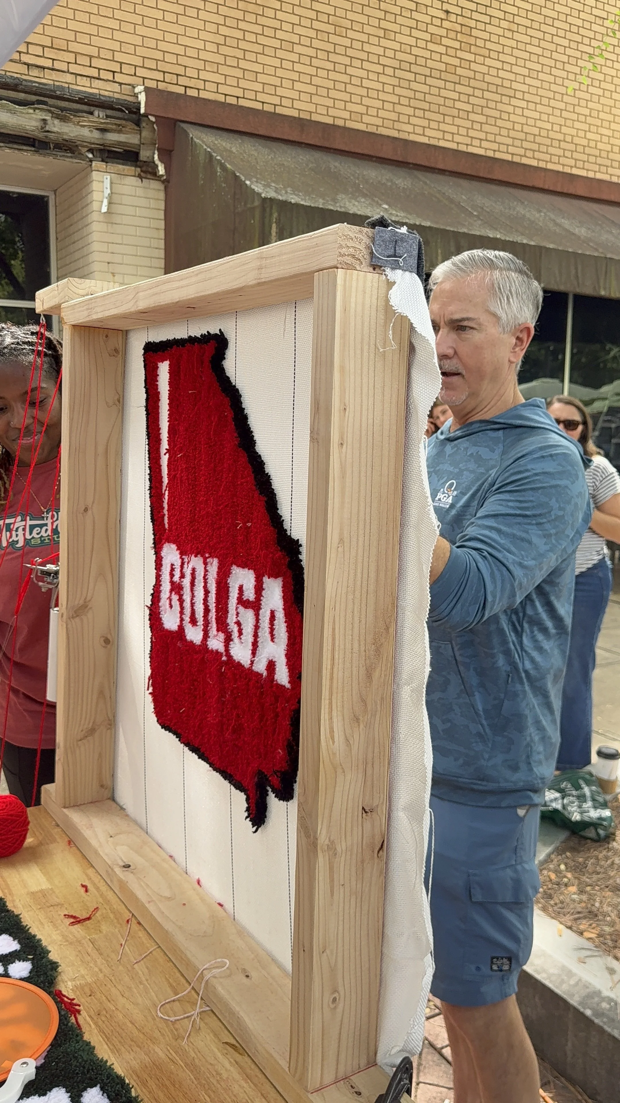 People working on a craft project involving a red and black embroidered patch for the state of Texas with the name 'GOLGA' on it, on a wooden frame outdoors.