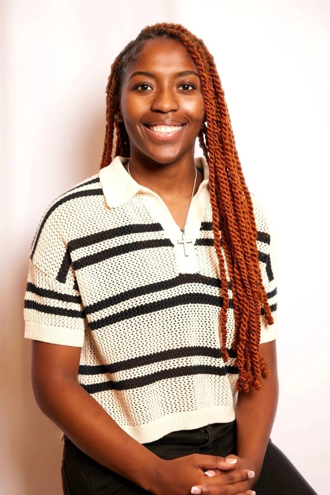 A young woman with long, reddish-brown dreadlocks smiling, wearing a white and black striped short-sleeve shirt and a cross necklace.