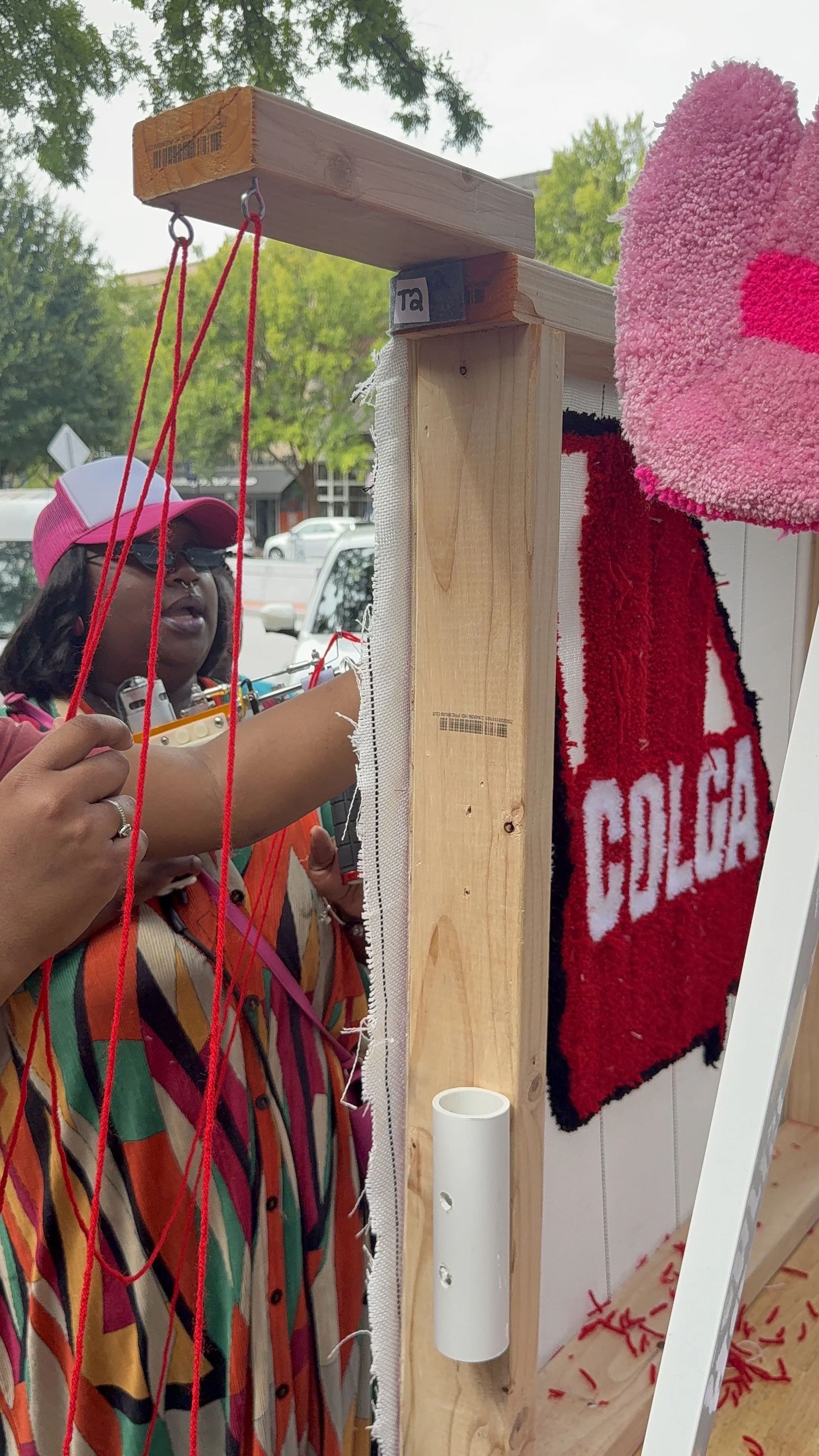 A woman wearing a pink cap and multicolored striped shirt stands behind a wooden structure with red strings hanging from it. There is a pink towel with red writing hanging on the right side of the structure, and some red strings are on the floor. The