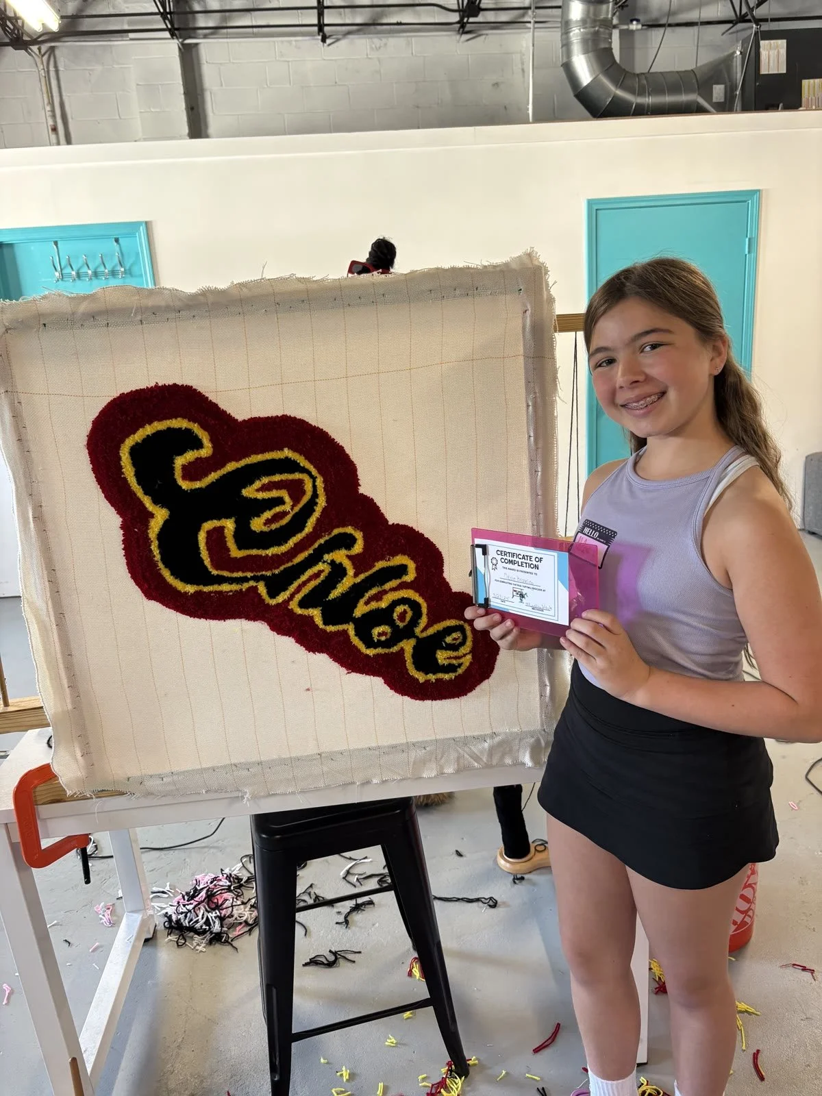 A girl holding a certificate of completion standing next to a hooked rug that says 'Smile' in black and yellow letters with a red background, in an indoor setting with a white wall, blue doors, and industrial ventilation.