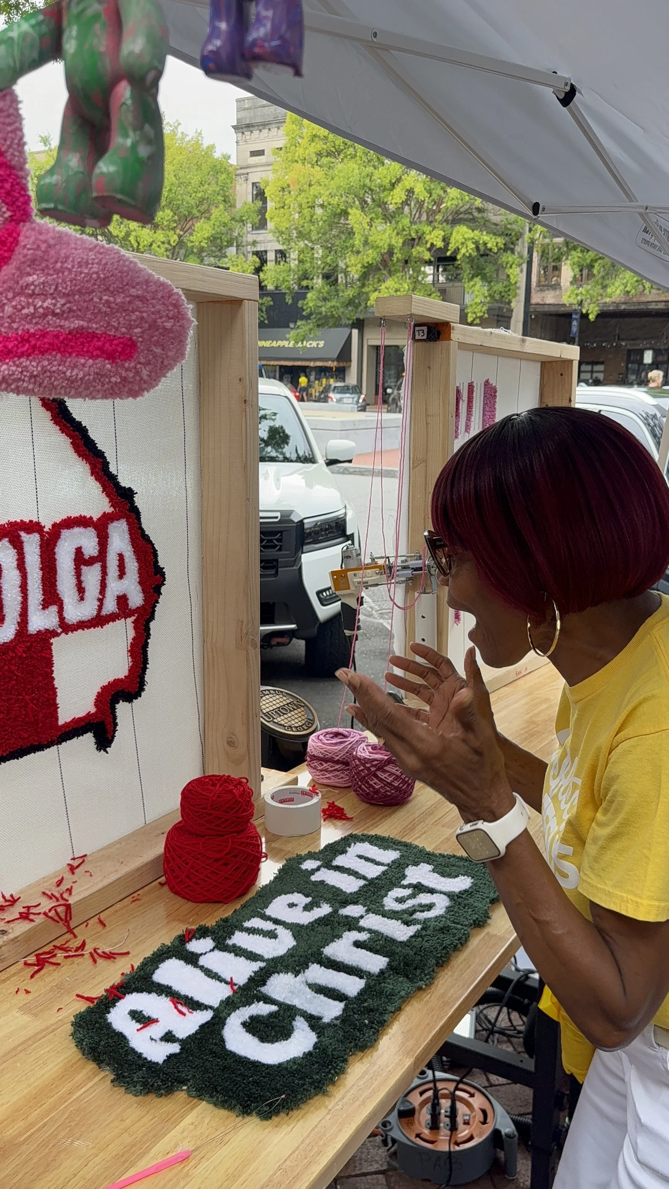 A woman with short red hair and glasses weaving a hemp or yarn banner at an outdoor craft booth. The banner has white and green text, with the visible portion reading "EDUCAS" and "ON". She is wearing a yellow T-shirt and has a white watch on her wri