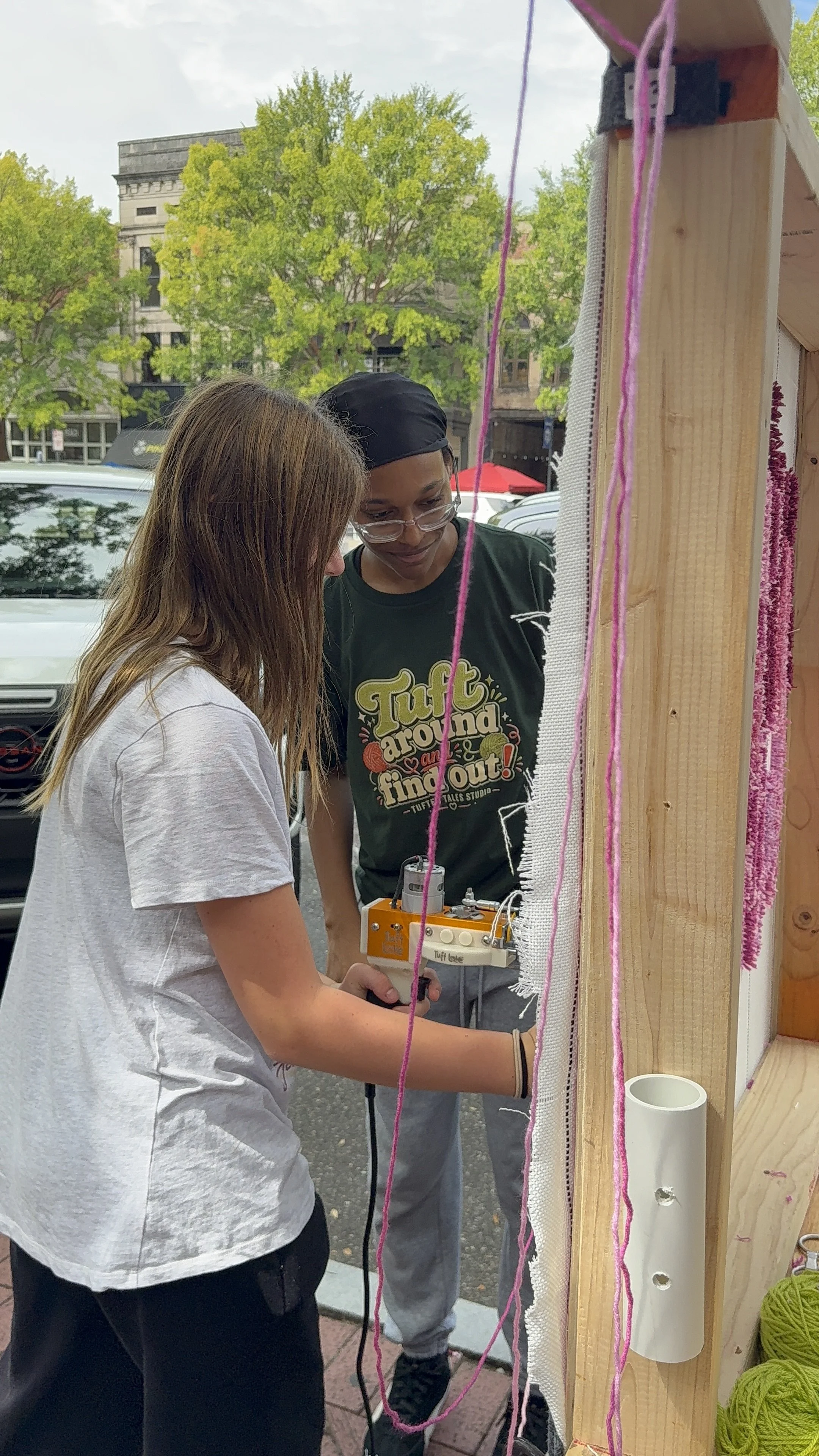 Two young women working on a yarn weaving project outdoors, with trees and buildings in the background.