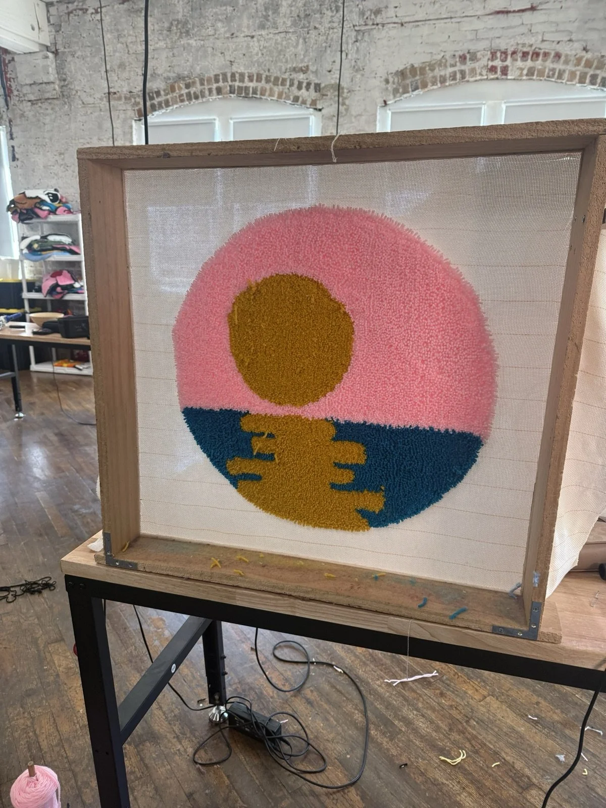 A circular artwork depicting a stylized sunset or sunrise over water, with pink sky, yellow sun, and dark blue water, mounted in a wooden frame with a mesh backing.