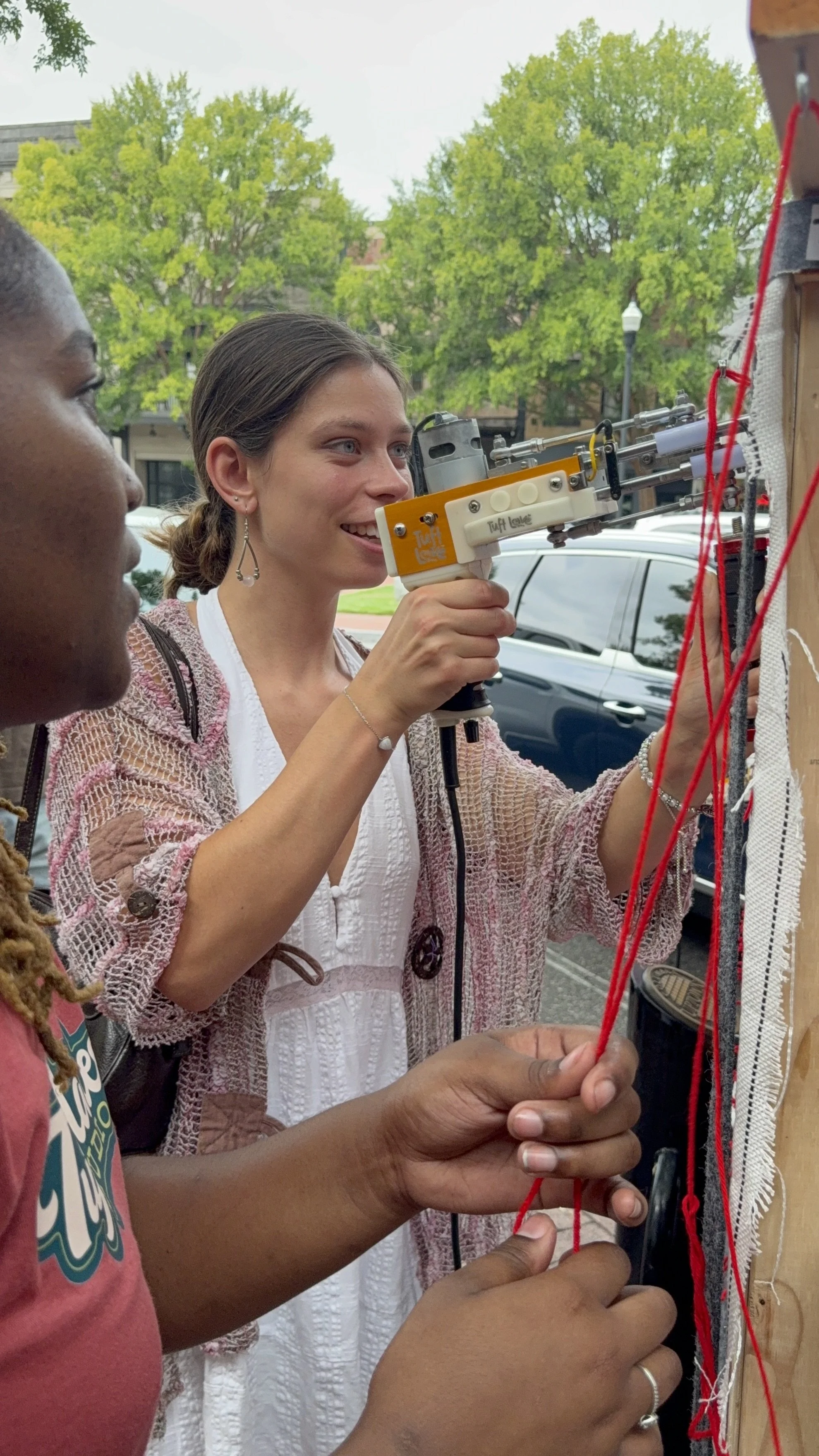 Two women working on a project together outdoors, with trees and a vehicle in the background. One woman is holding a tool and looking at a wall with red thread and string, while the other woman is observing.