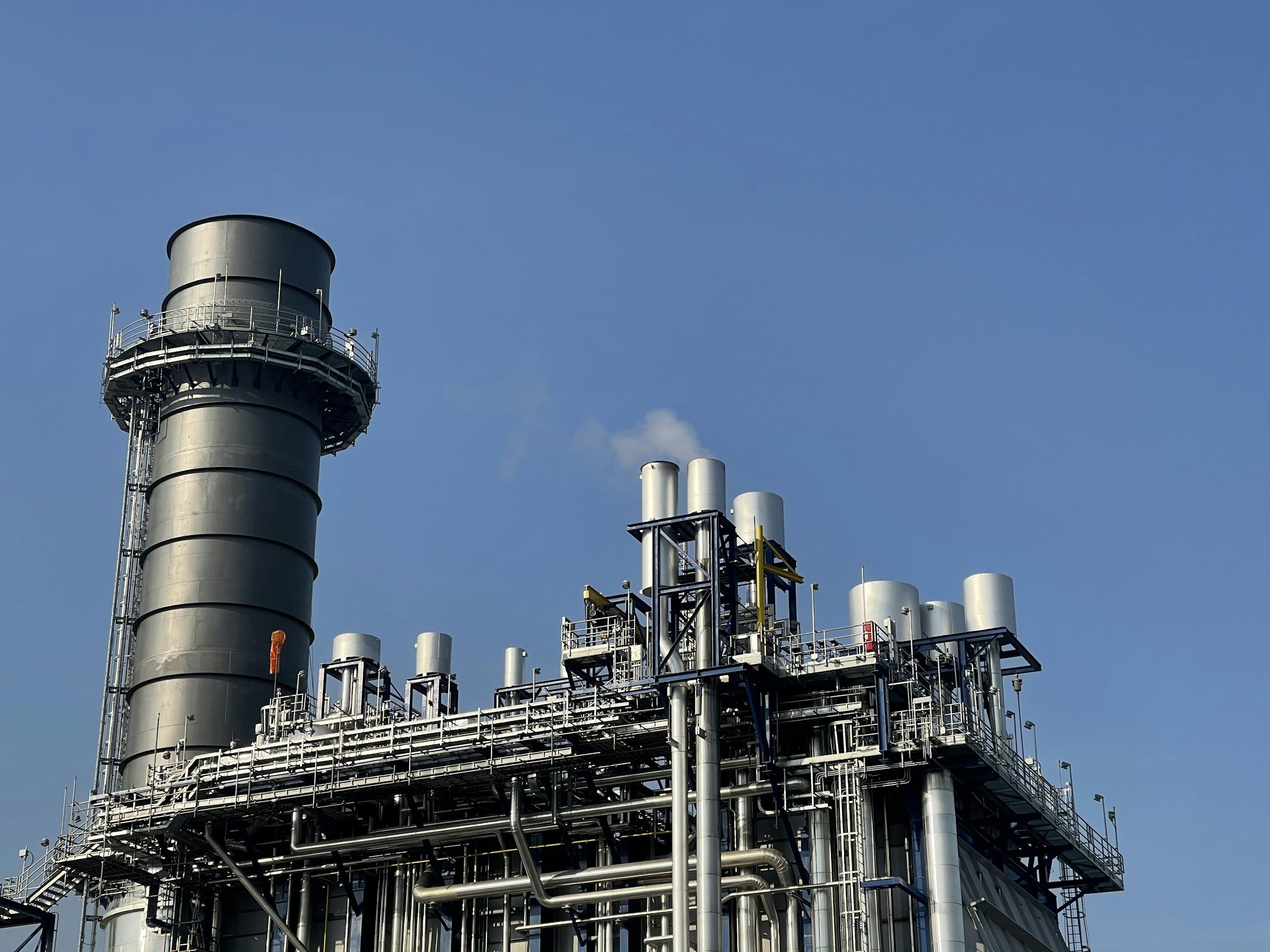 Industrial factory with a large metallic chimney and complex piping system under a clear blue sky.