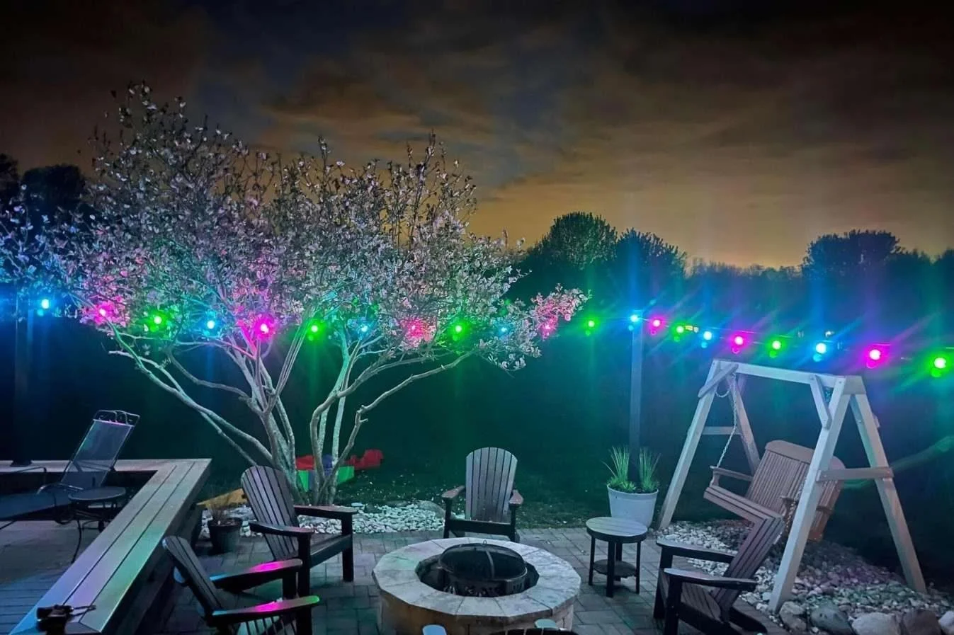 Nighttime backyard scene with a lit blooming tree, colorful string lights, wooden swing, Adirondack chairs around a fire pit, and a potted plant on a patio.