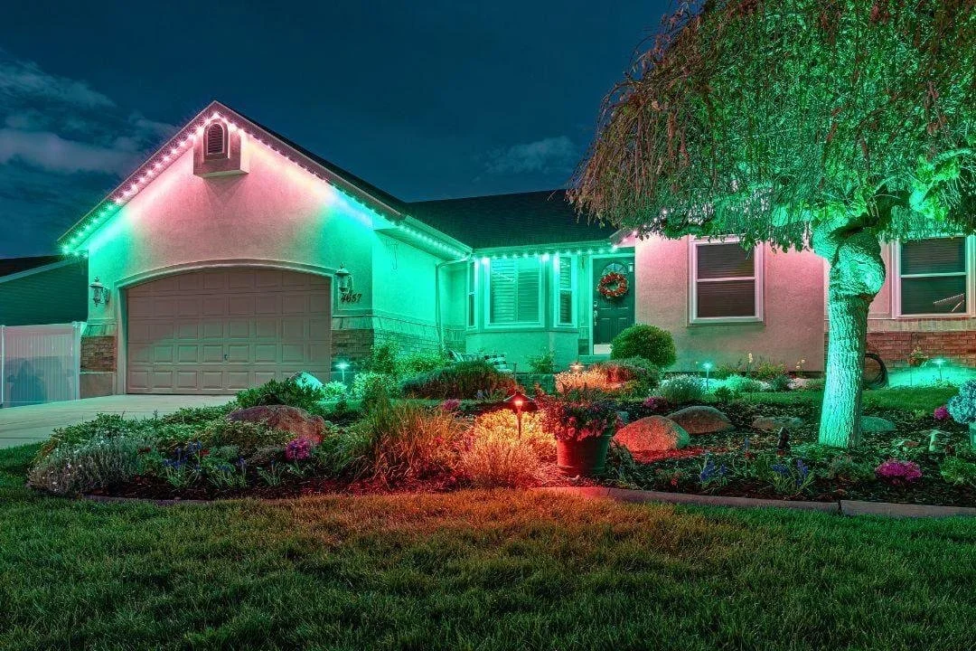 A house decorated with colorful Christmas lights at night, featuring green, red, and pink lighting on the exterior and on the trees and garden in the front yard.