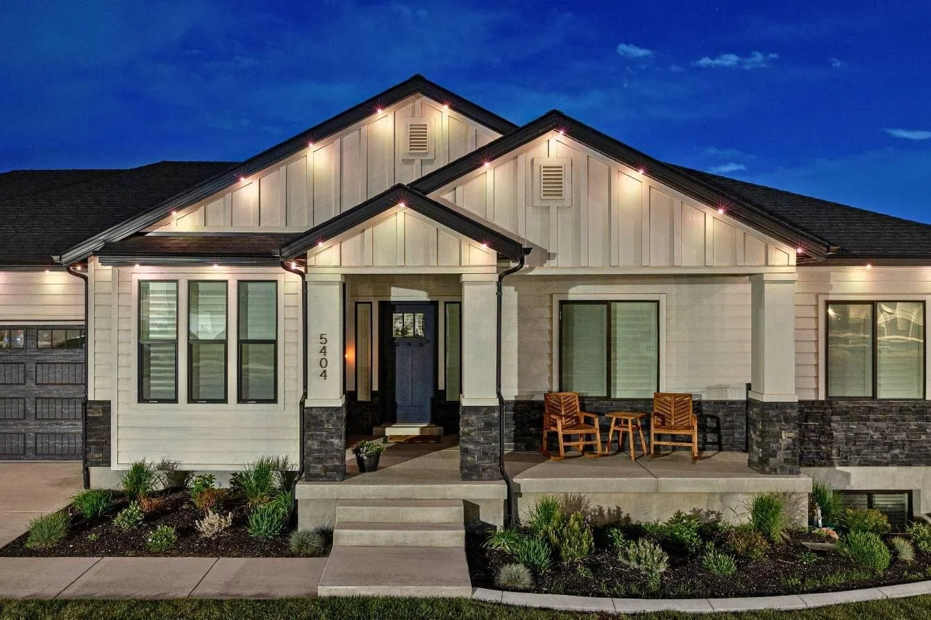 A modern house with a covered front porch, illuminated by exterior lights at dusk. The house has white siding, black window frames, and a black front door. There are two wooden chairs and a table on the porch, with landscaping in front of the house.