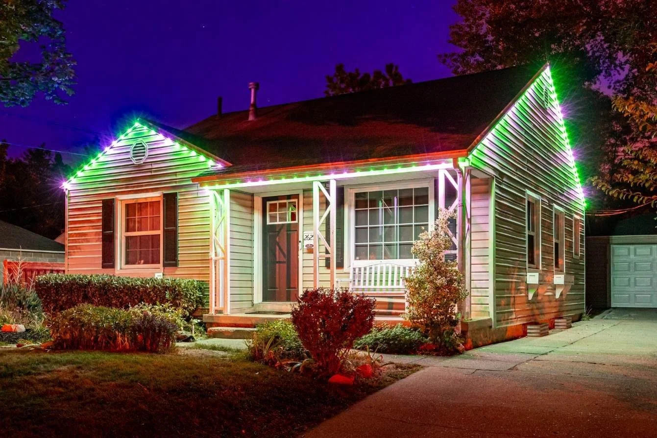 A house illuminated with colorful Christmas lights at night, featuring green, red, and purple lights outlining the roof and sides, with a front yard and driveway.