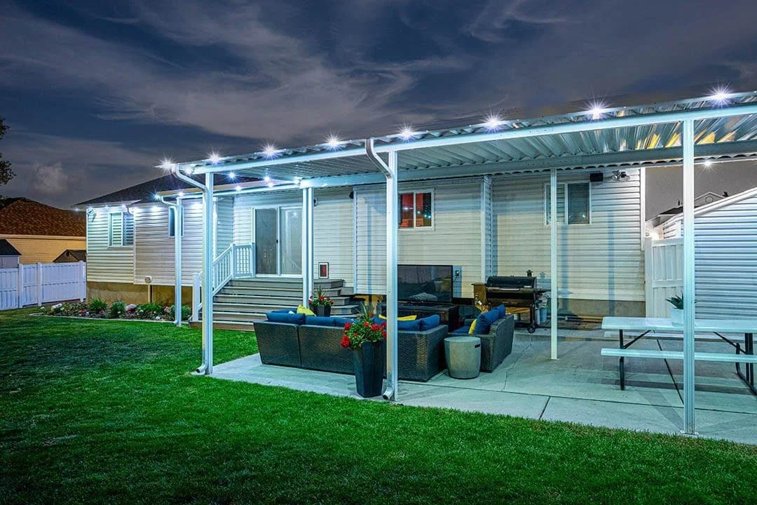 Backyard patio at night with string lights, outdoor furniture, a grill, and a picnic table under a covered patio attached to a house with siding.