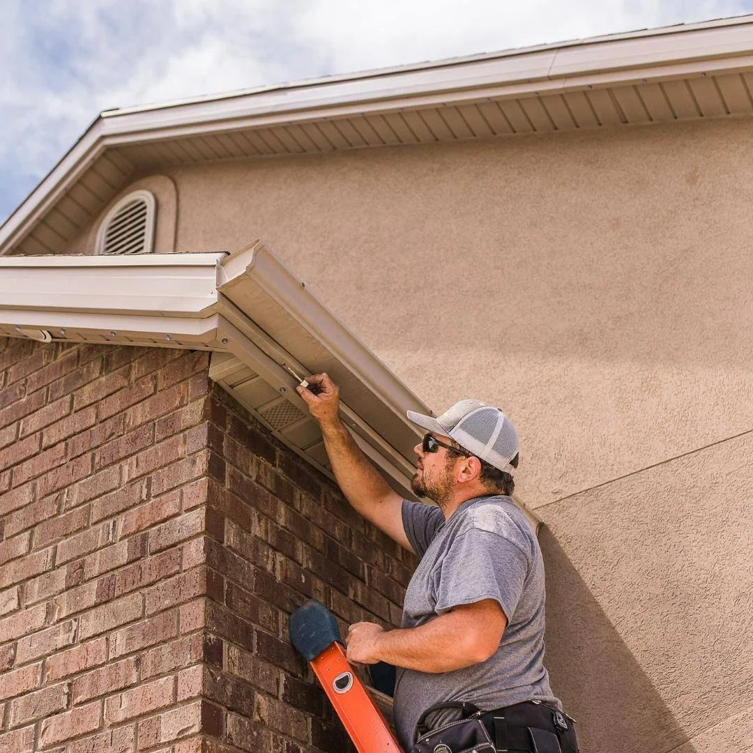 A man with a gray t-shirt, sunglasses, and a baseball cap working on the gutter of a house, standing on a ladder.