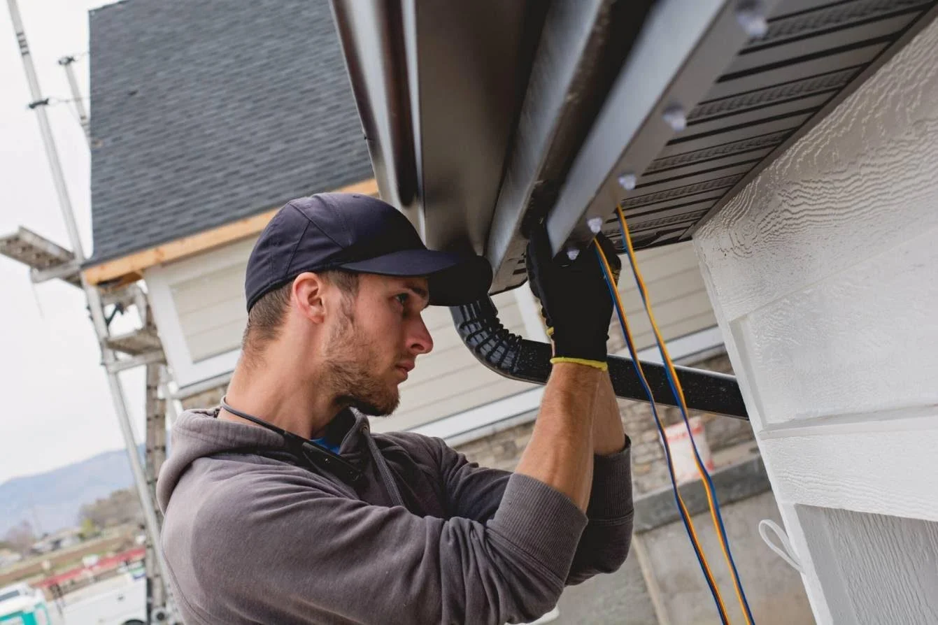 A man wearing a black cap and gray hoodie working on electrical wiring underneath an HVAC unit outside a house.
