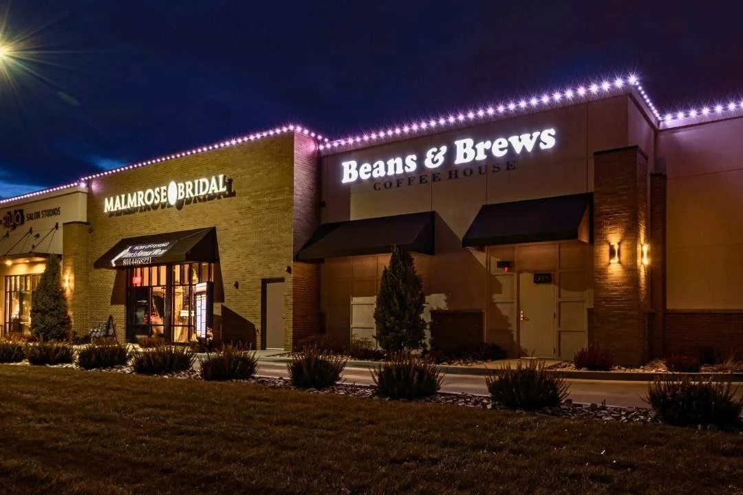 Night view of a shopping plaza with illuminated storefronts including Malmrose Bridal and Beans & Brews Coffee House, with small shrubs and trees in front.