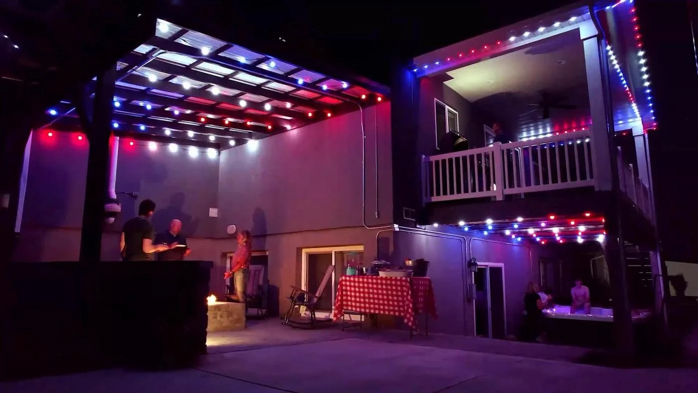People gathered on a patio decorated with red, white, and blue string lights, with a hot tub, a grill, and a table with a red checkered tablecloth at night.