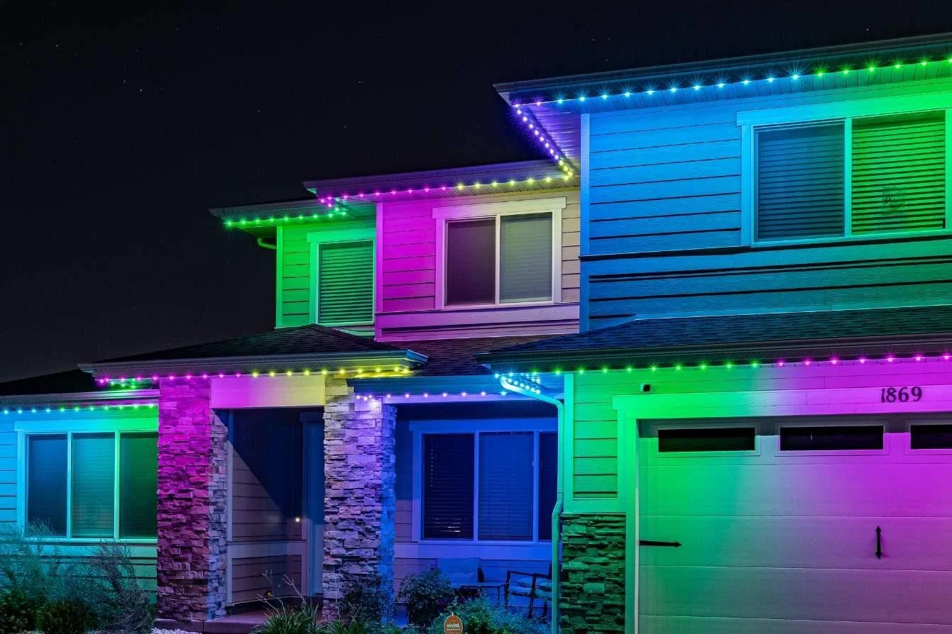 A two-story house decorated with colorful Christmas lights at night, with green, purple, and blue lights outlining the roof and windows.