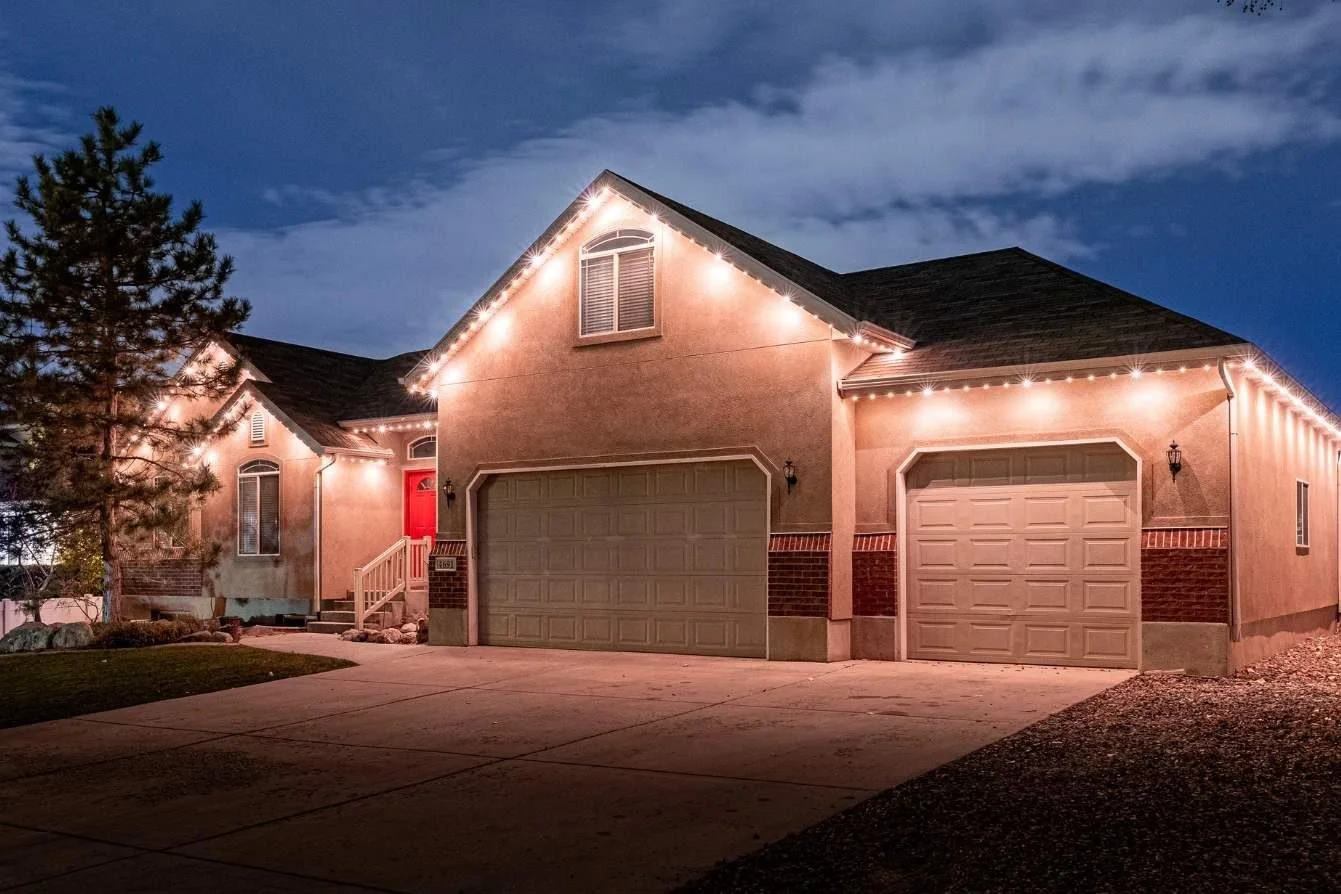 A two-story house at dusk with exterior lights and string lights outlining the roof, a front porch with stairs, a red front door, two garage doors, a tree on the left, and a driveway leading to the garages.