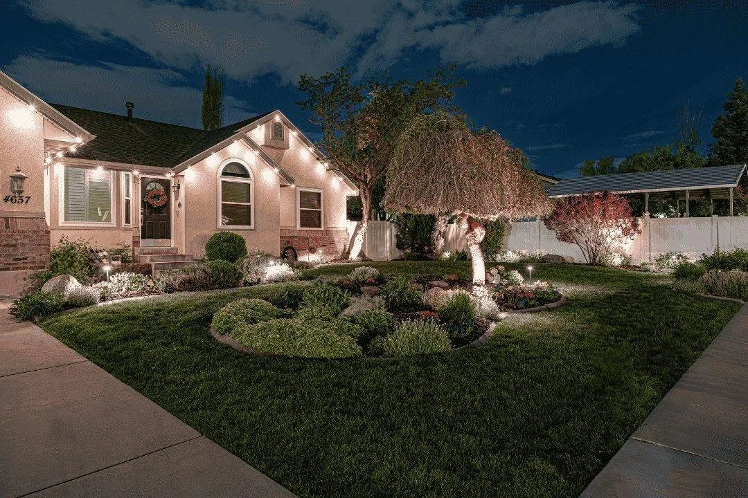 Nighttime view of a well-lit front yard with landscaped garden, trees, and a house decorated with string lights.