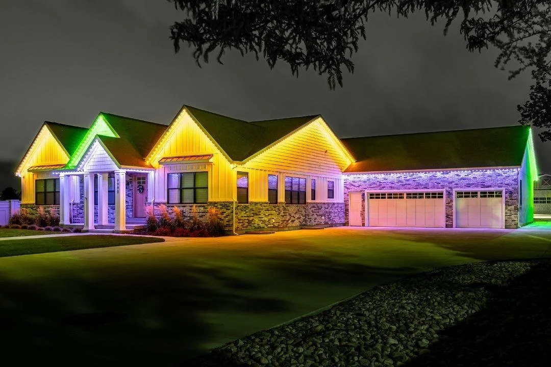 A house illuminated with colorful Christmas lights at night, featuring yellow, blue, purple, and green lights on the exterior. The house has a brick lower wall, white siding, and a three-car garage.