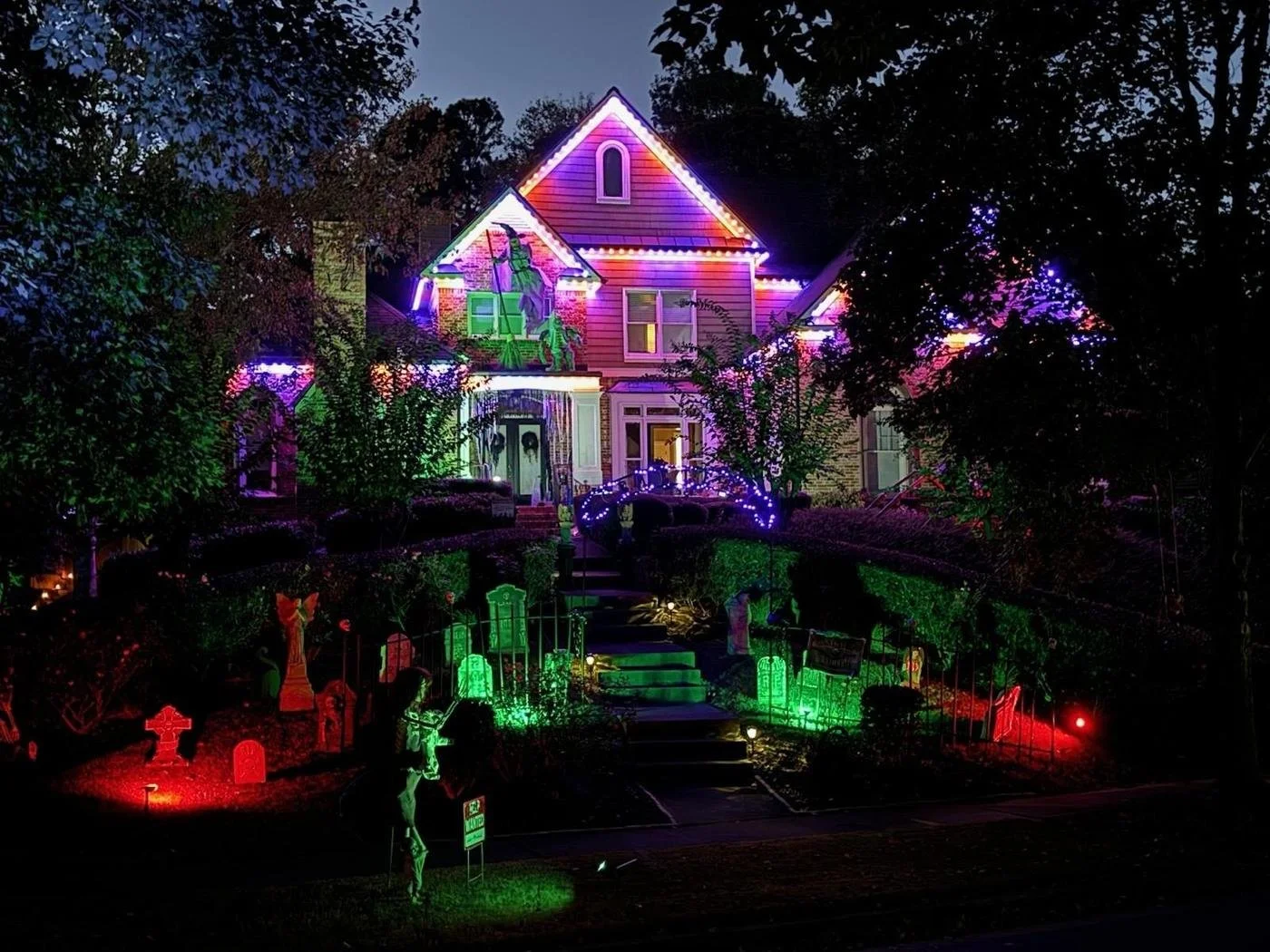 A house decorated with colorful Christmas lights at night. The house has multiple stories with a gabled roof, and the yard is illuminated with red, green, purple, and white lights. There are Halloween-themed decorations, including tombstones and a sk