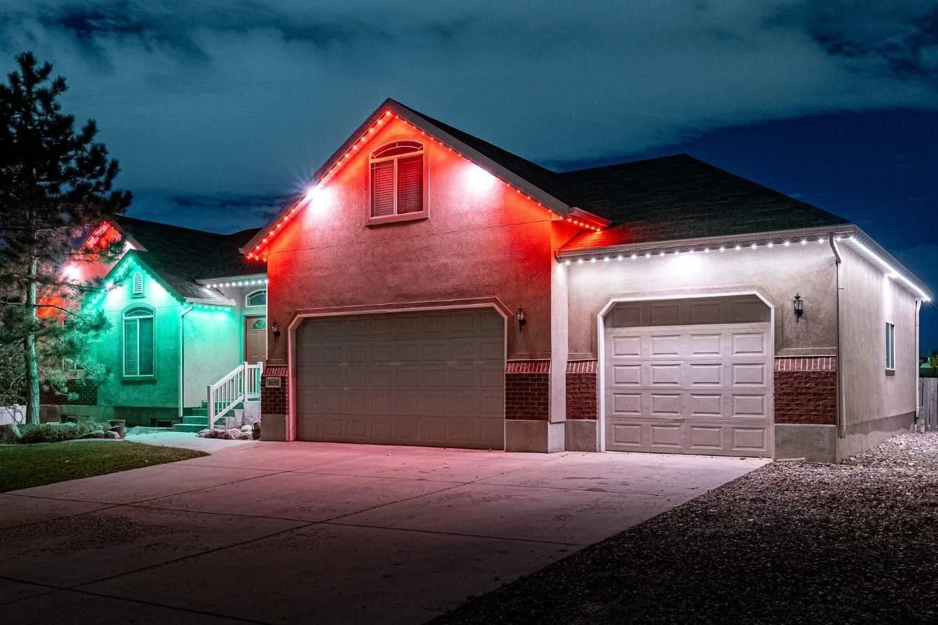 A house decorated with multicolored Christmas lights at night, featuring green, red, and white lights outlining the roof and garage doors.
