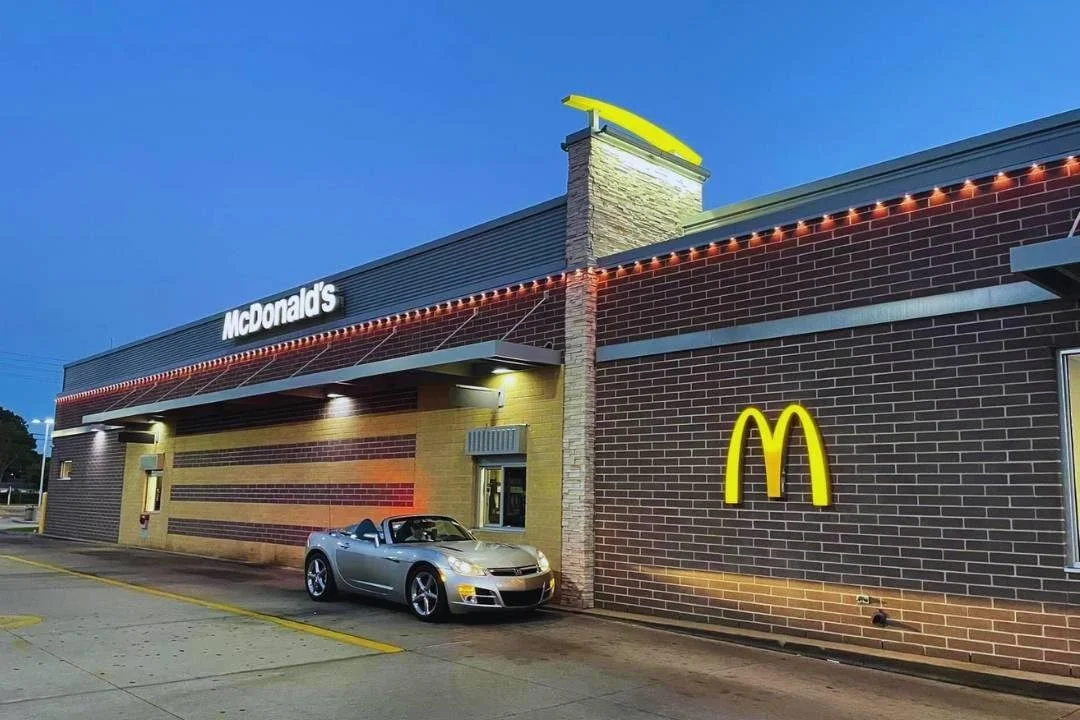 Exterior of a McDonald's restaurant at dusk with a yellow illuminated McDonald's logo, a convertible car parked in front, brick walls, and string lights along the roofline.