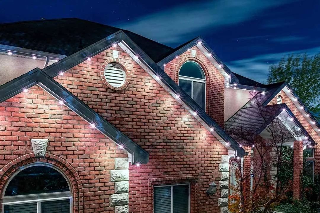 Nighttime view of a brick house with exterior string lights outlining the rooflines, arched windows, and decorative stone accents.