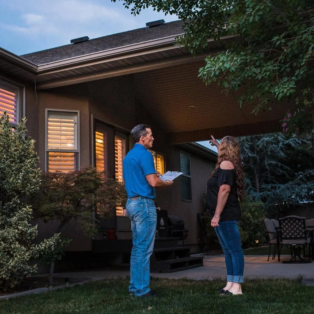 A man in a blue shirt holding a clipboard converses with a woman in black on a backyard patio at dusk, near a house with lit windows.