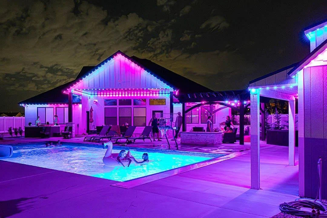 Nighttime scene of a backyard with a lit swimming pool shaped like a swan, surrounded by lounge chairs and a house decorated with colorful string lights.