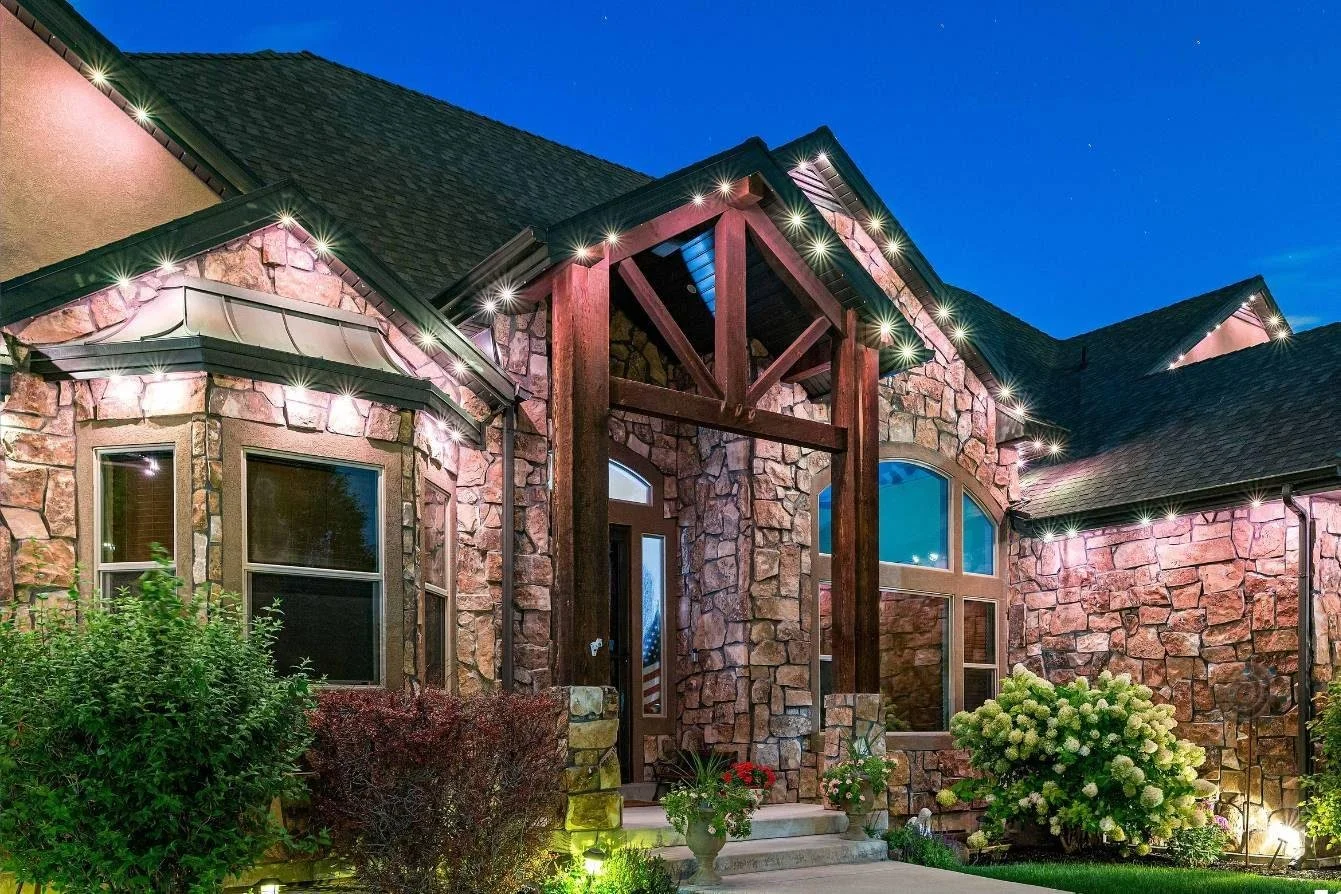 A stone house with a peaked roof at night, illuminated by exterior lights, featuring large arched windows and a wooden entryway with steps, surrounded by landscaped bushes and flowers.