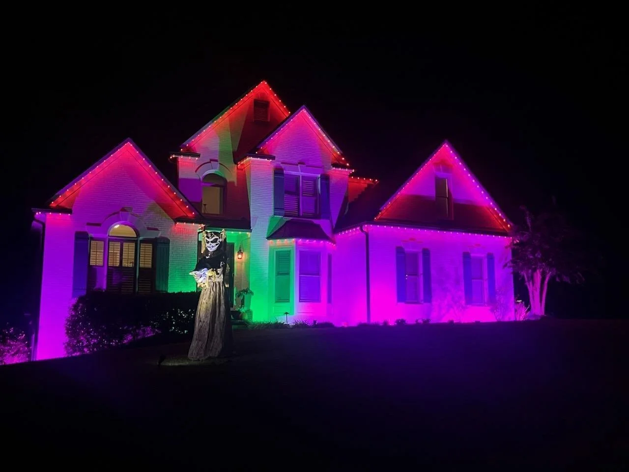 A house decorated with colorful Halloween lights at night, with a spooky figure dressed as a witch or skeleton standing in front.