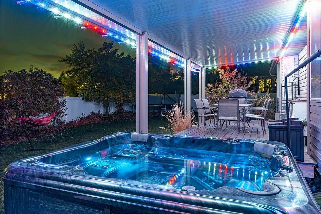 Backyard with illuminated hot tub, wooden deck with patio table and chairs, string lights, hammock, and trees at night
