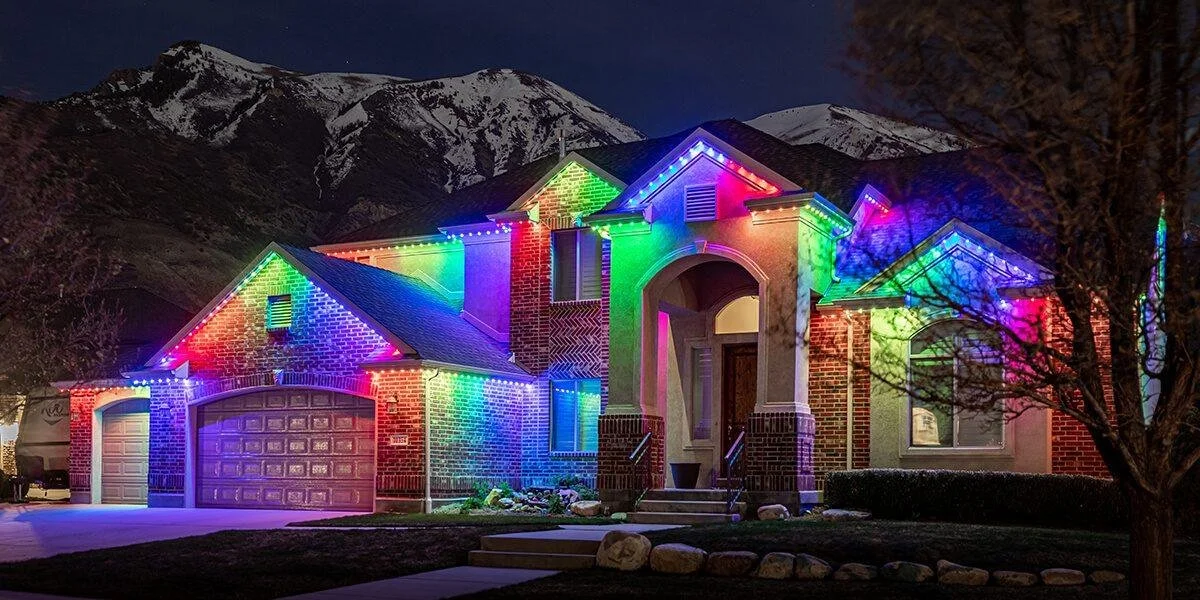 A two-story house decorated with colorful Christmas lights at night, with snow-covered mountains in the background.