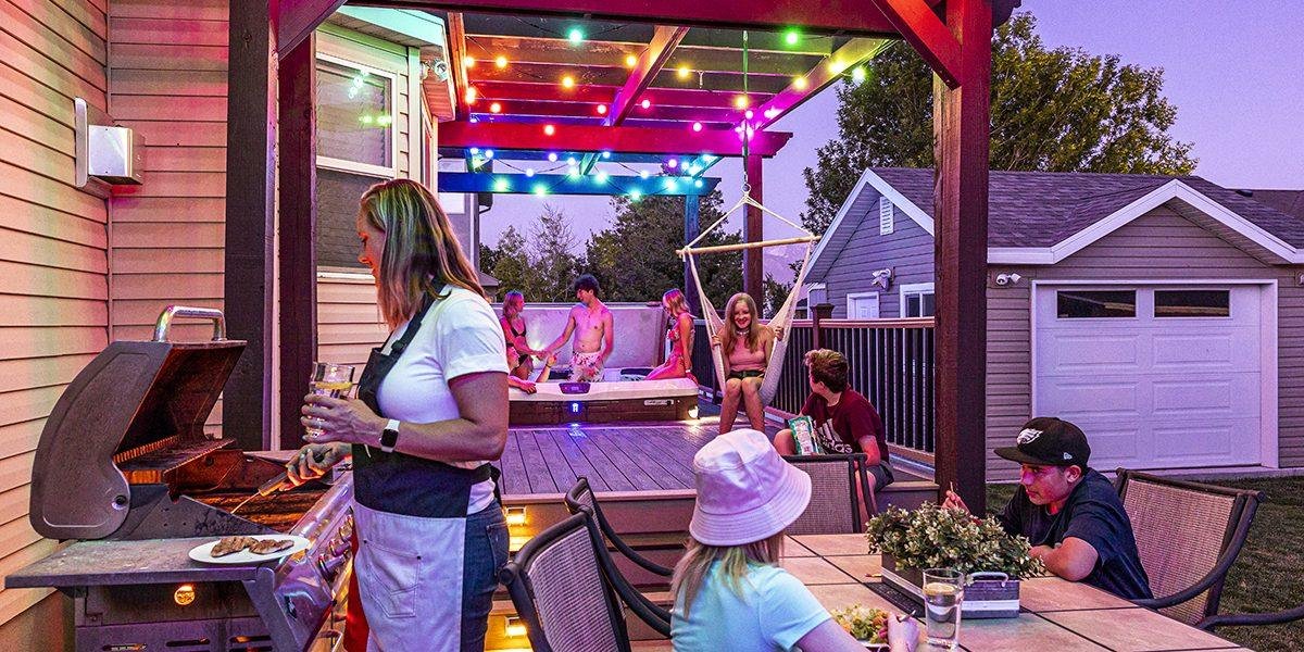 People enjoying a backyard party with a barbecue grill, a hot tub, and colorful string lights, with a woman and children relaxing at a table and others playing in the background.