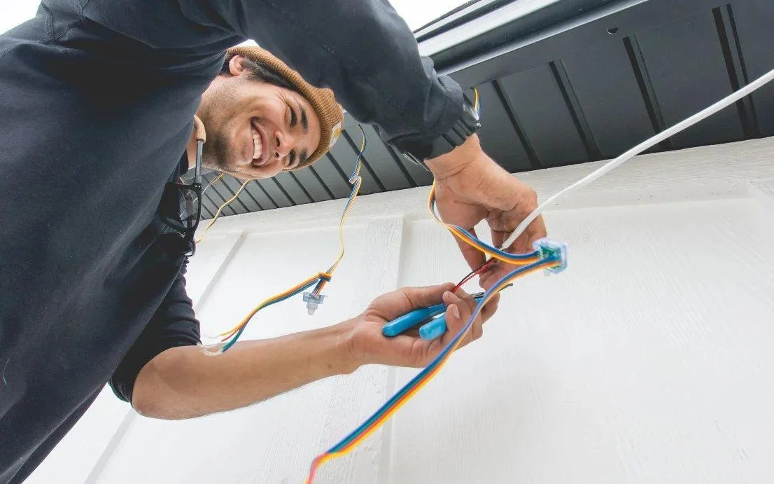 A man smiling while working on electrical wiring inside a building, with various wires and tools.
