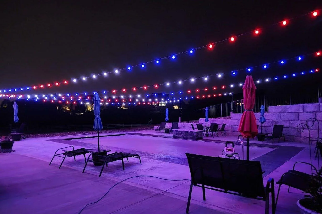 Nighttime outdoor patio with string lights in red, white, and blue, purple lighting on the ground, and closed umbrellas and chairs scattered around.