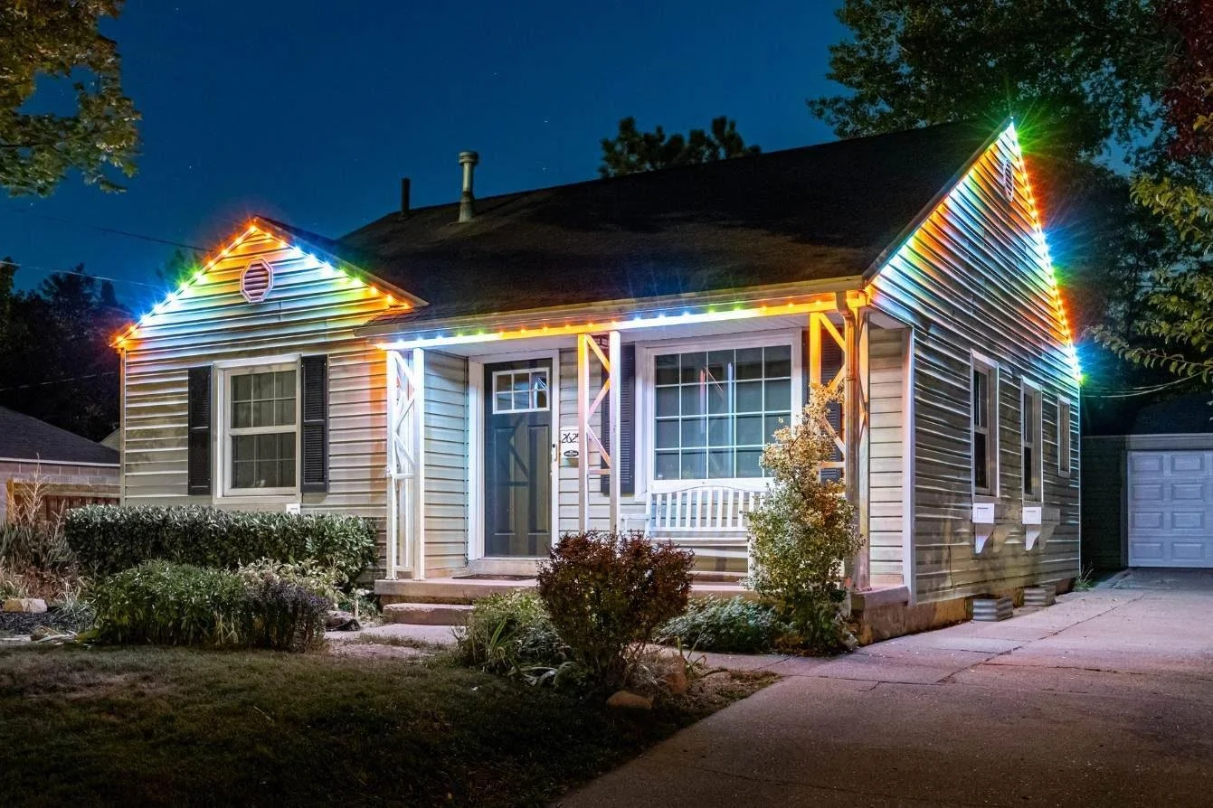 A house decorated with colorful Christmas lights at night, with a dark sky and trees in the background.